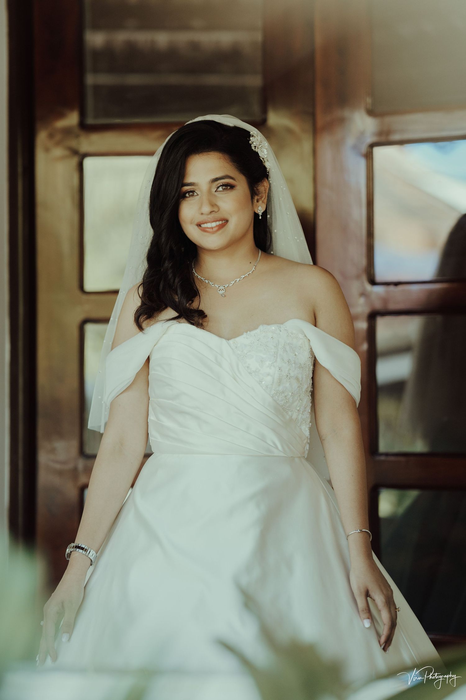 A bride in an elegant off-shoulder white wedding gown with embroidered bodice and veil poses inside a vintage setting.