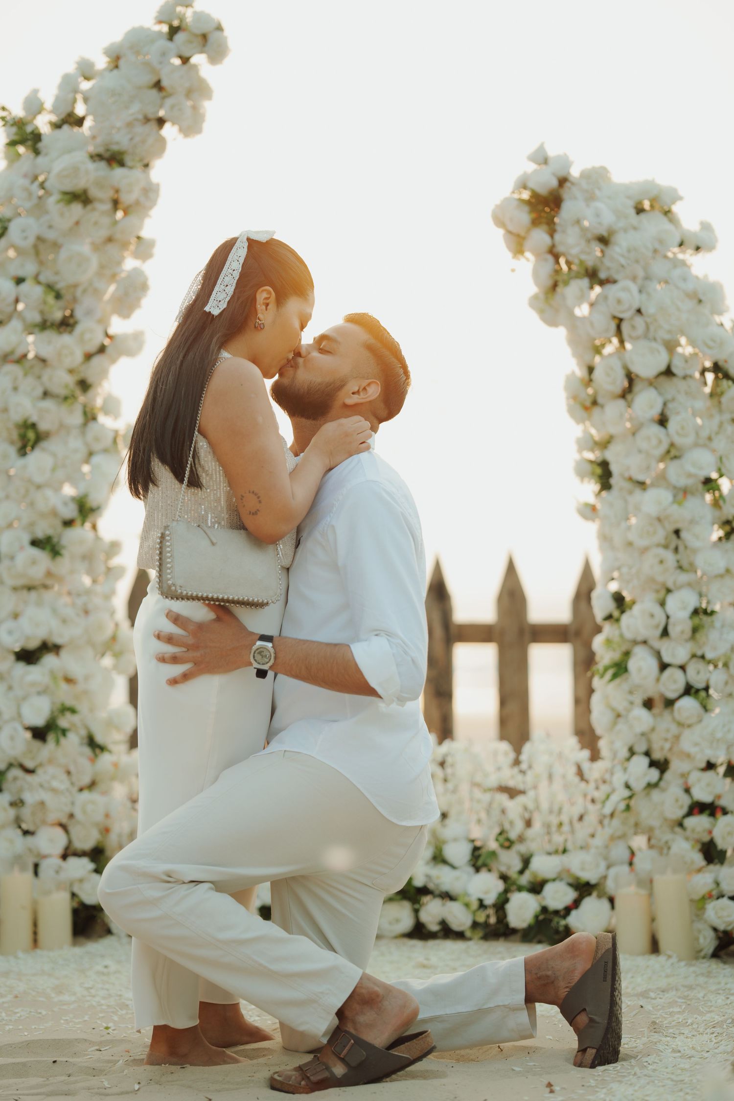 Couple sharing a romantic kiss during a wedding proposal surrounded by white floral arches on a beach.
