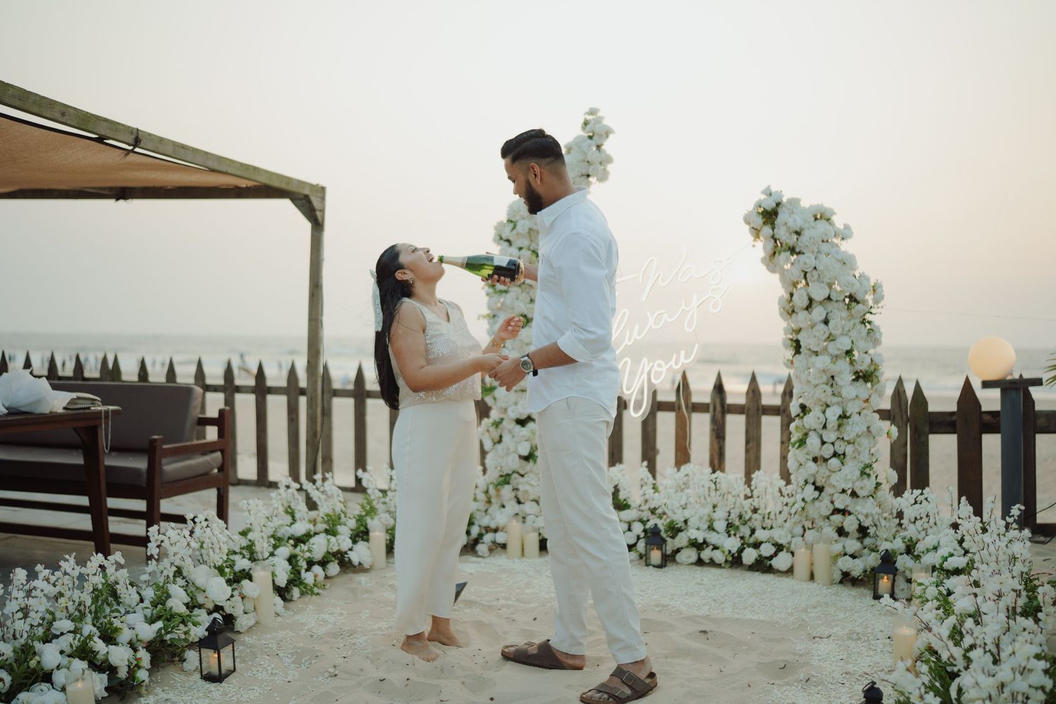 Couple sharing a romantic moment at a floral beach setup with white flowers and arch at sunset.