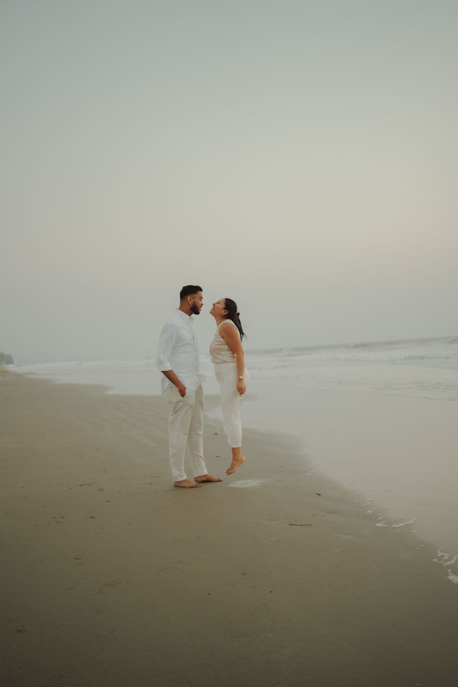 A couple in matching white outfits standing barefoot on a misty beach, gazing at each other romantically.