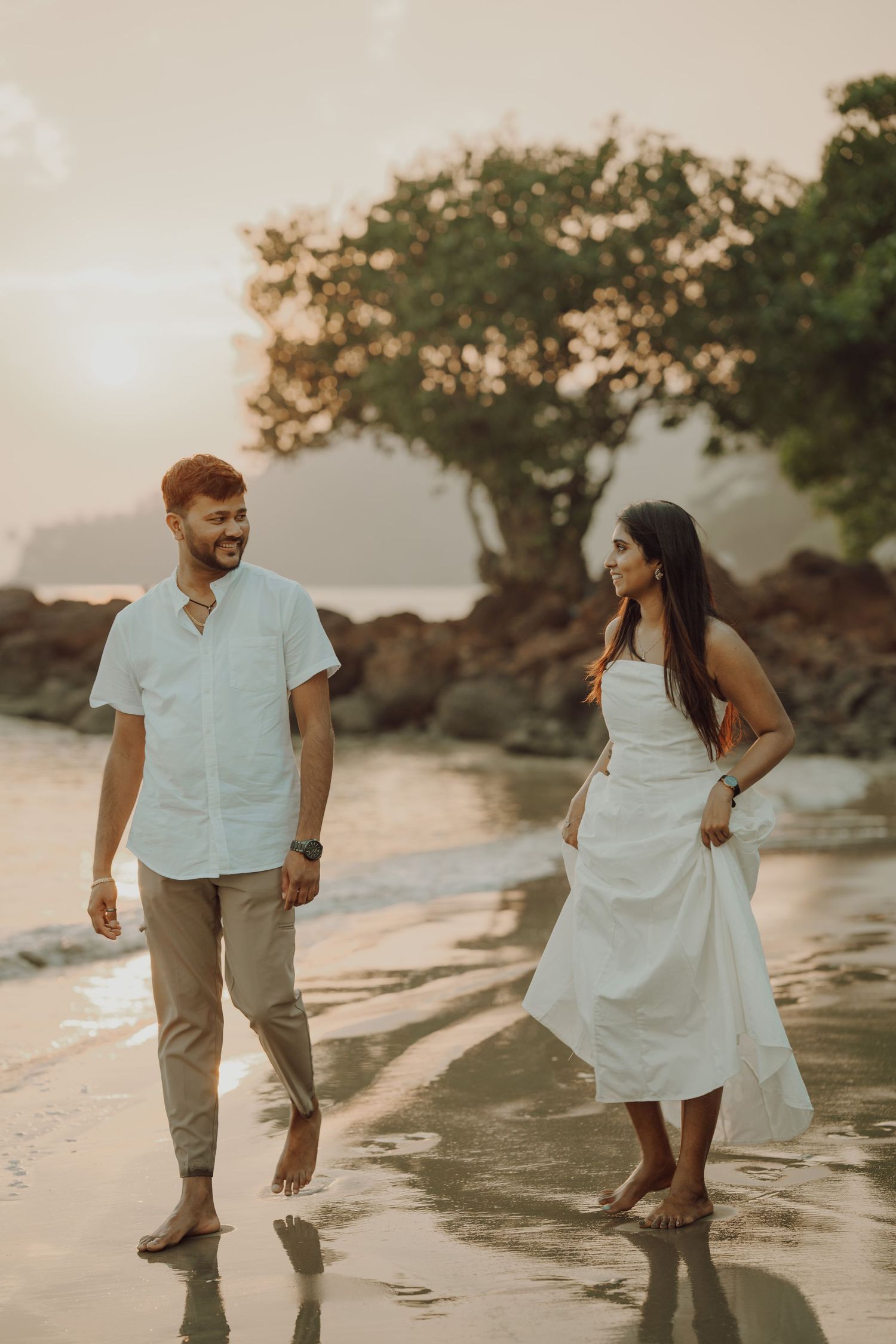 Couple in white and beige outfits walking playfully together on a beach at sunset.