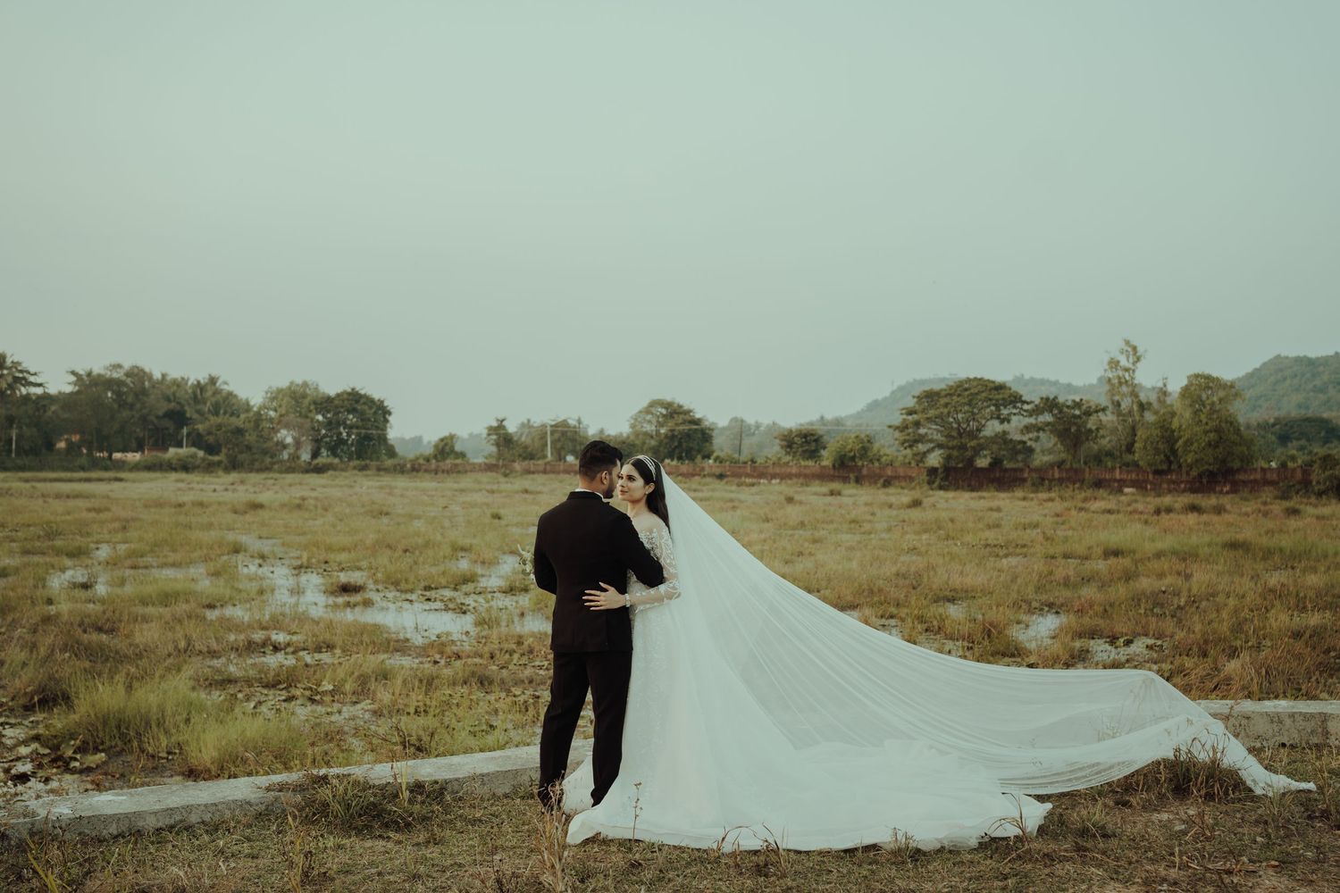 A dramatic wedding photo with long flowing veil against an overcast landscape.