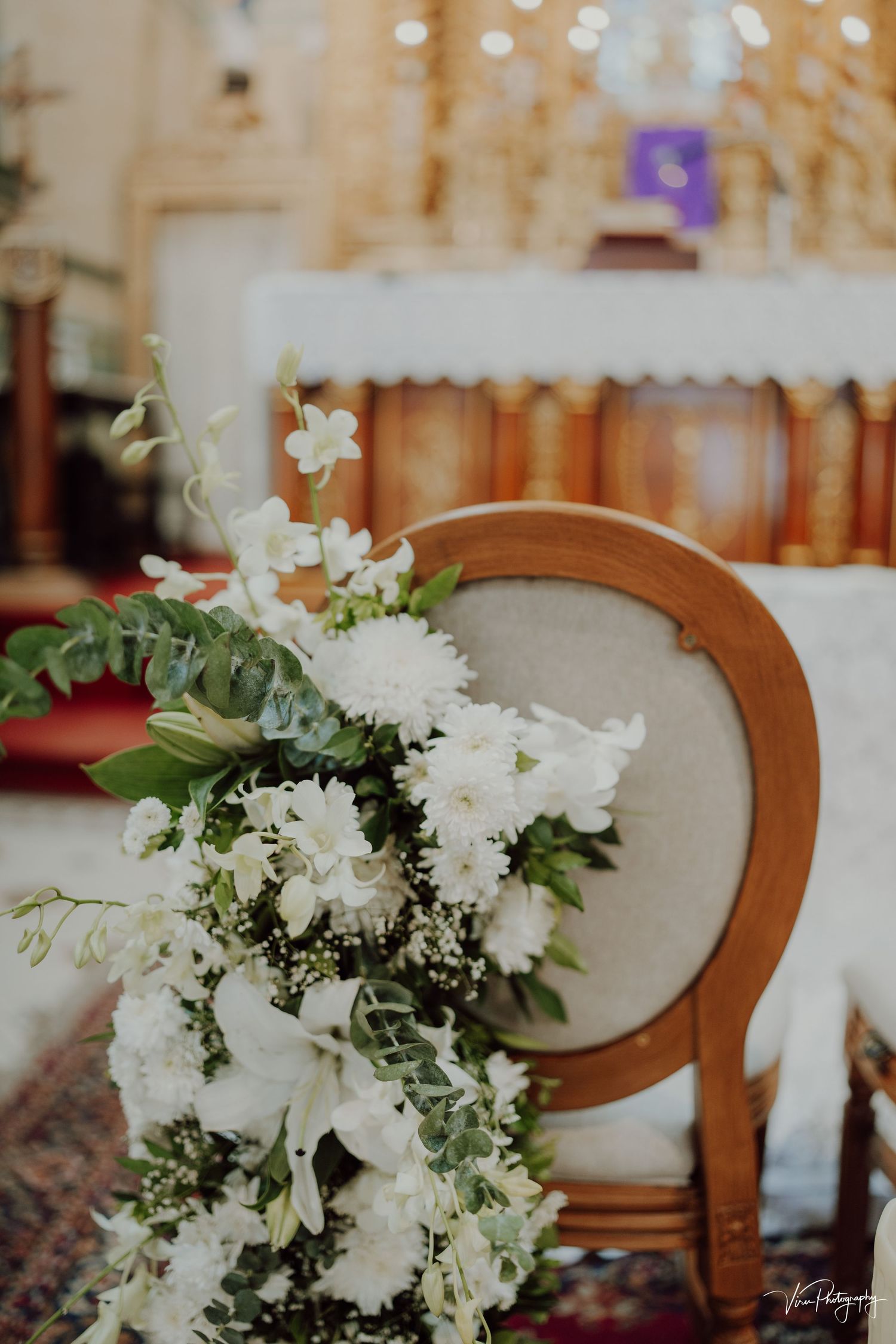 Close-up of elegant white floral arrangement with greenery resting on an antique upholstered chair at a wedding ceremony.