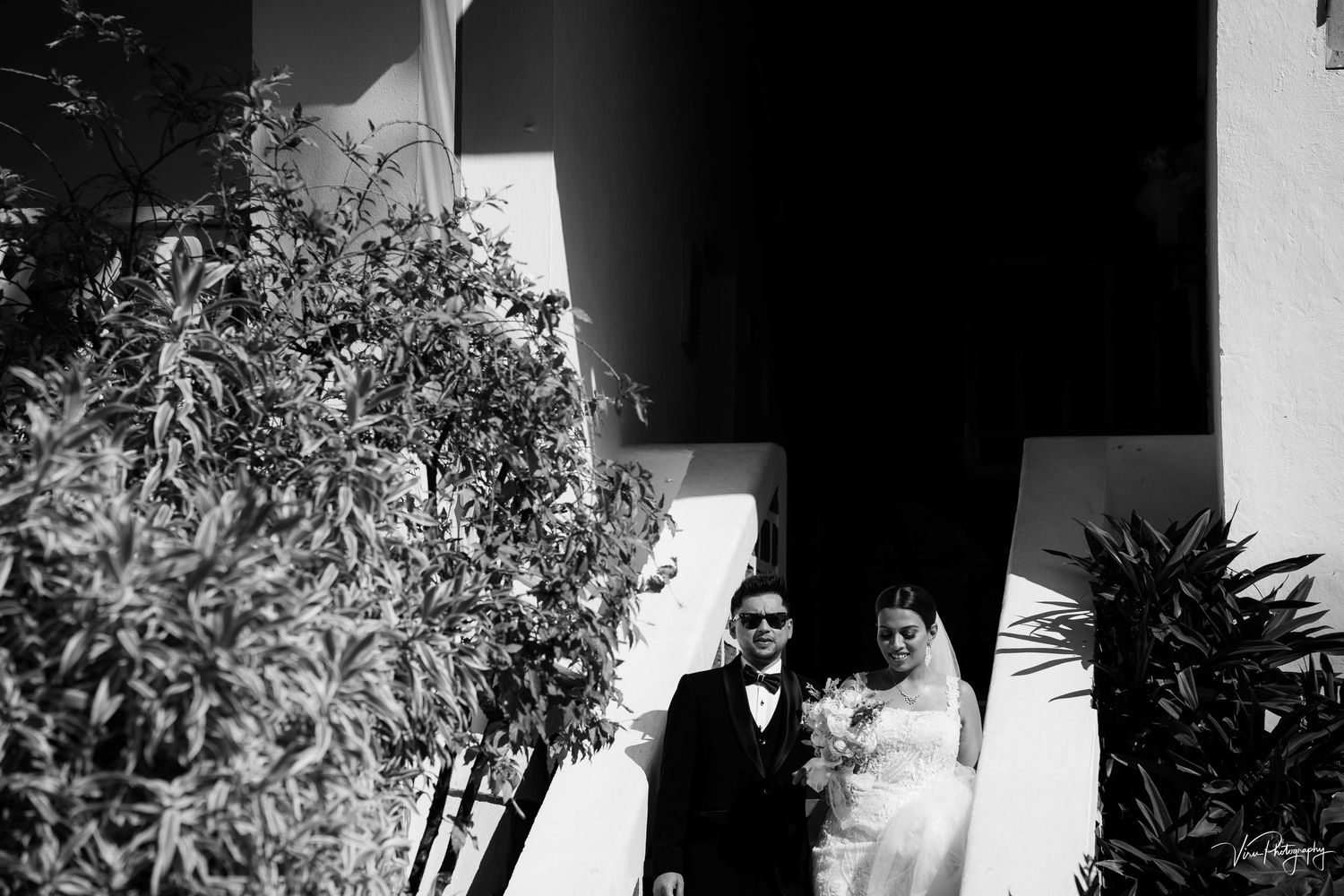 A black and white artistic wedding portrait against a white villa backdrop with tropical foliage.