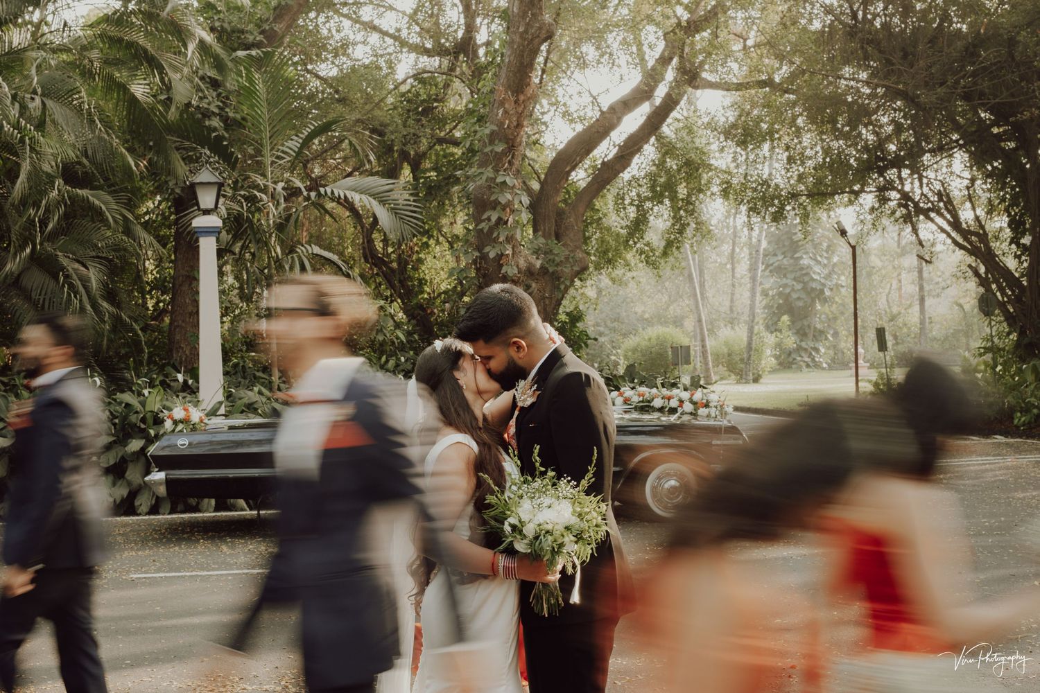A couple shares an intimate moment during their wedding ceremony while guests blur past in the foreground in a garden setting.