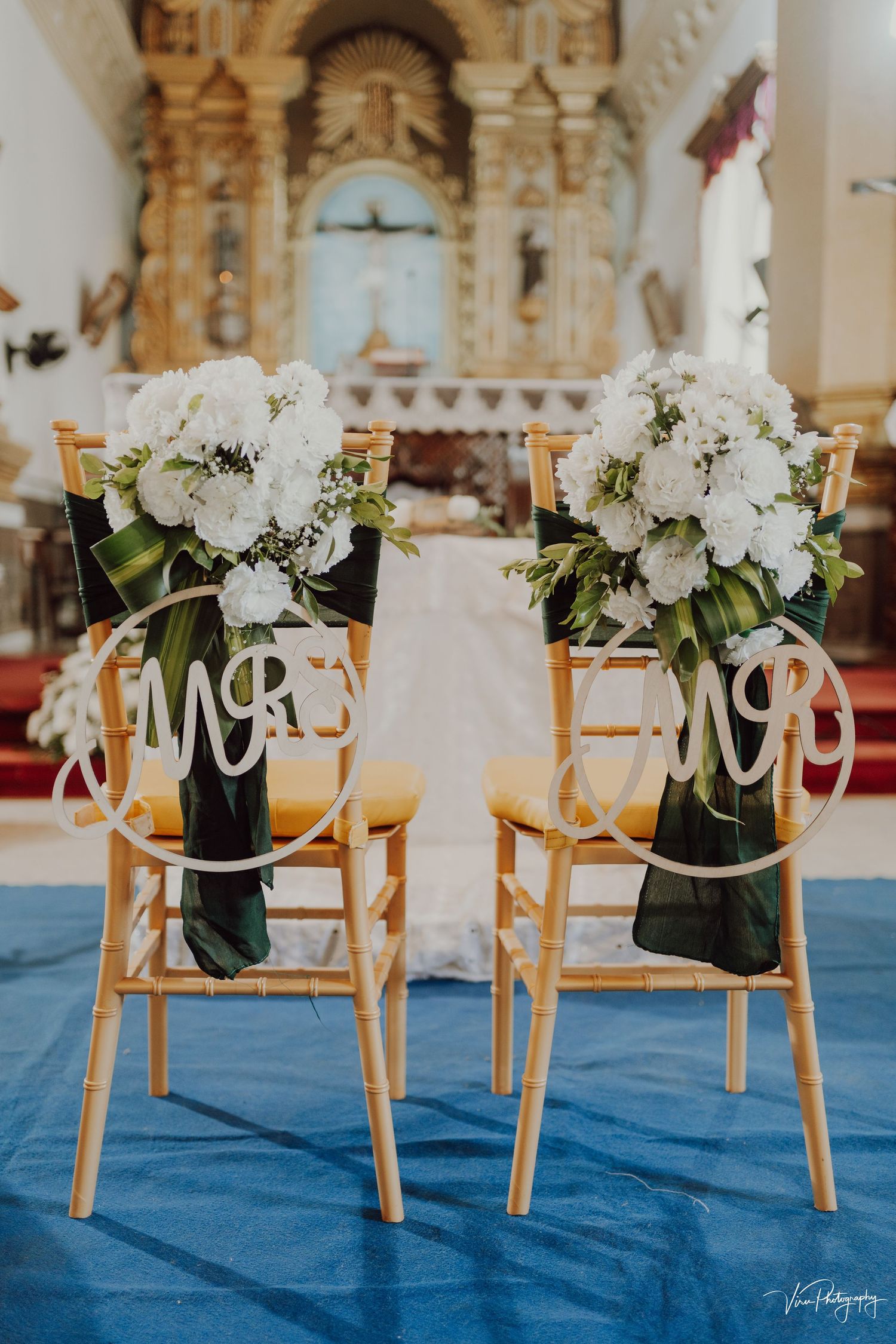 Two wooden chairs with white floral bouquets and 'Mr' signs displayed at a church wedding ceremony on blue carpet.