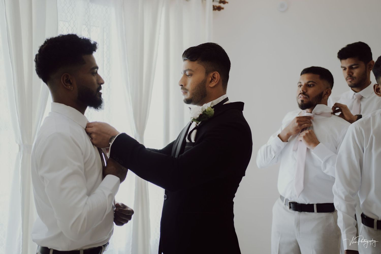 A groomsman helps adjust the tie of another member of the wedding party as they prepare in a bright white room.