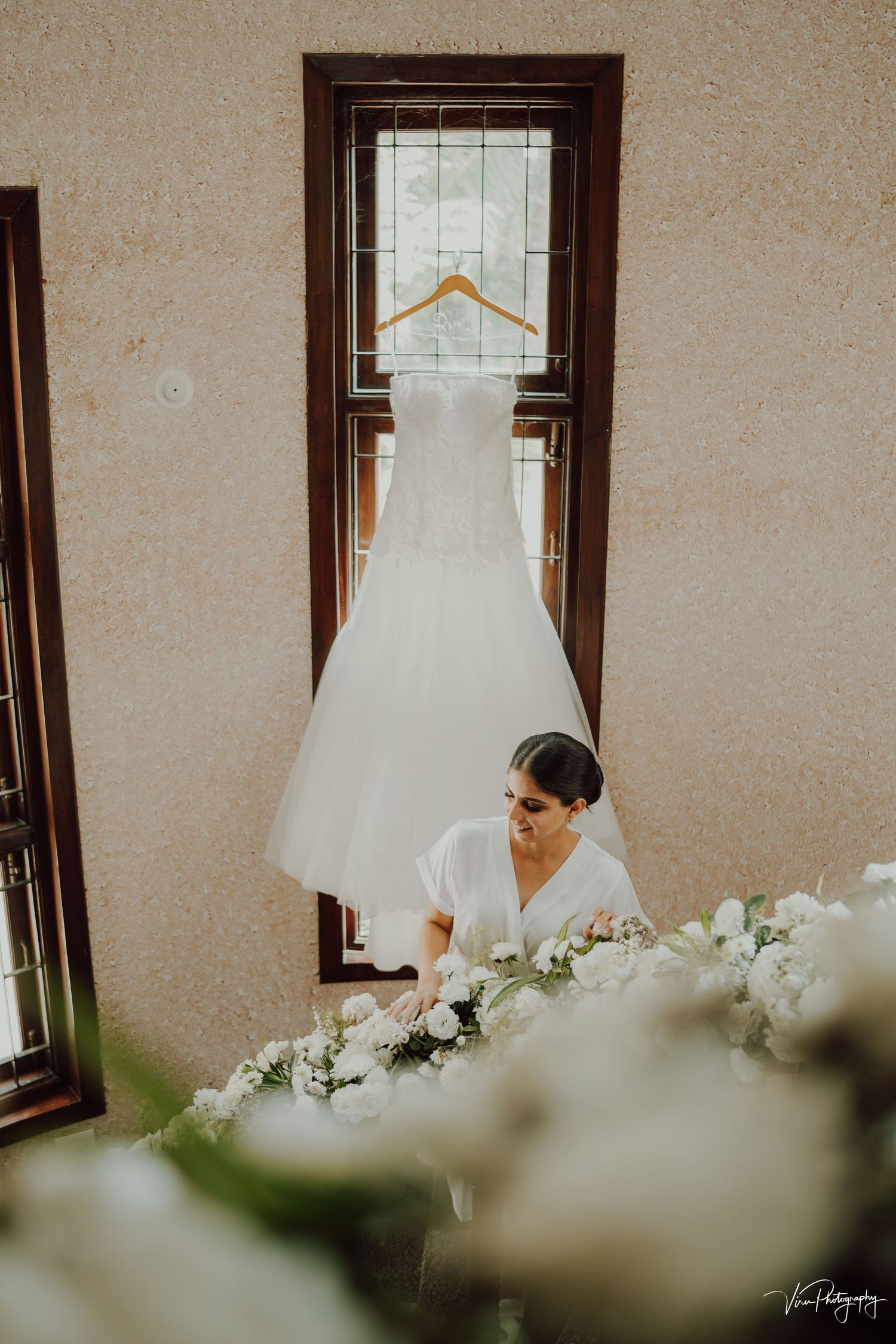 A white wedding dress hangs in a window as flowers frame a bridal preparation scene in warm natural lighting.