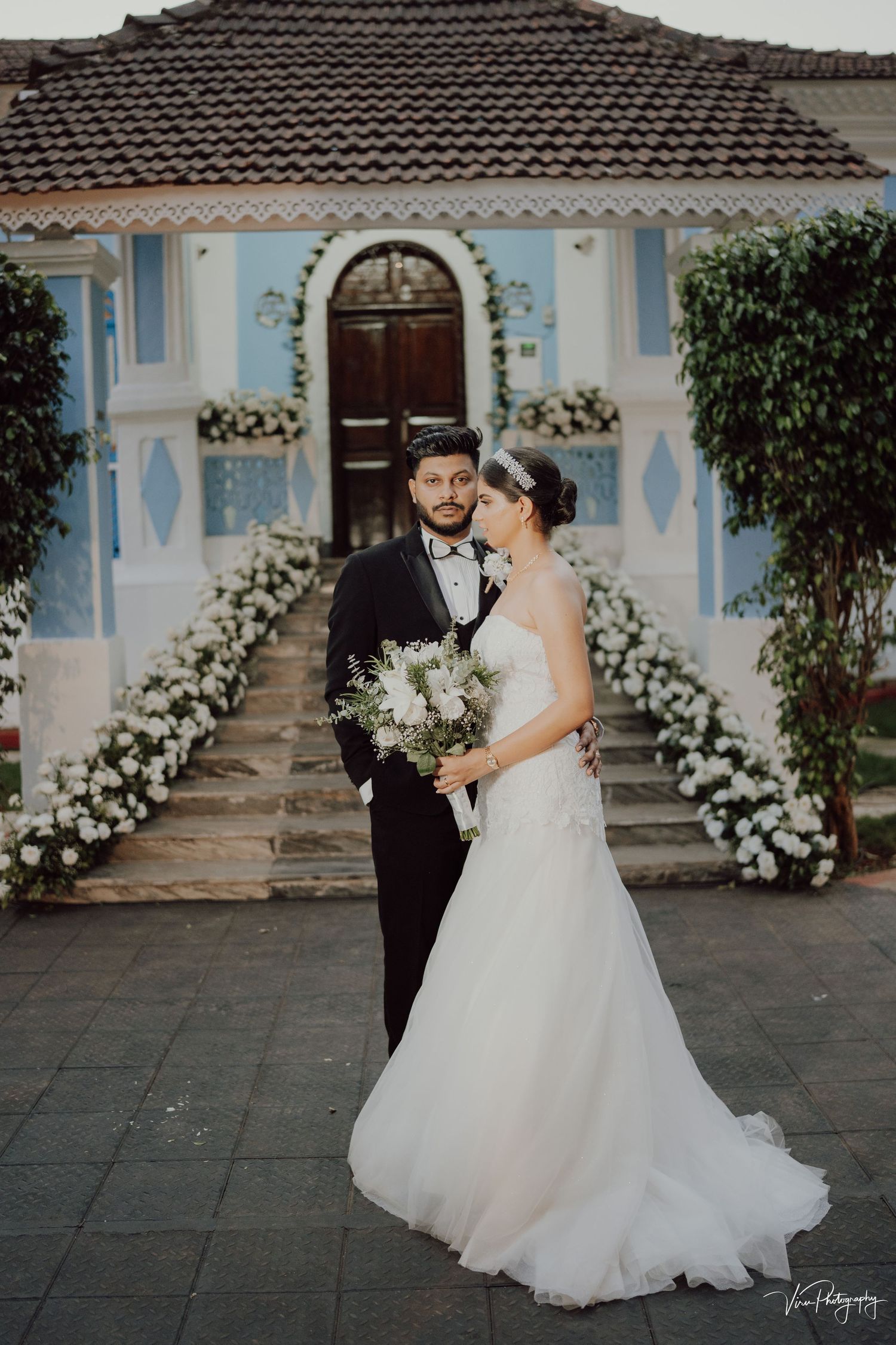 Wedding couple poses in front of blue building with arched doorway and white floral decorations.