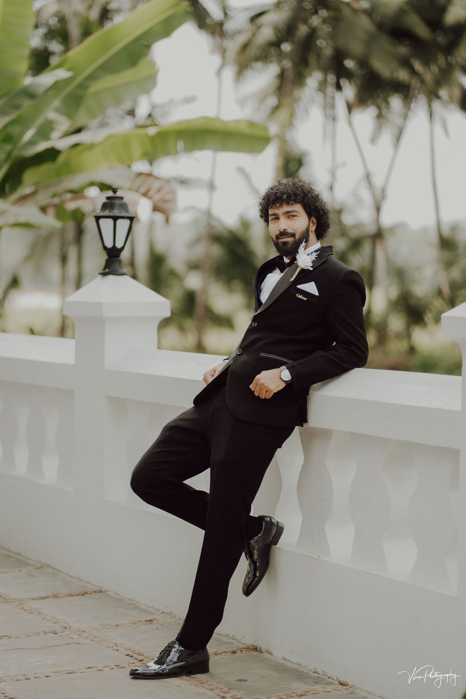 A groom in a classic black tuxedo leans casually against a white balustrade with palm trees and a lantern in the background.