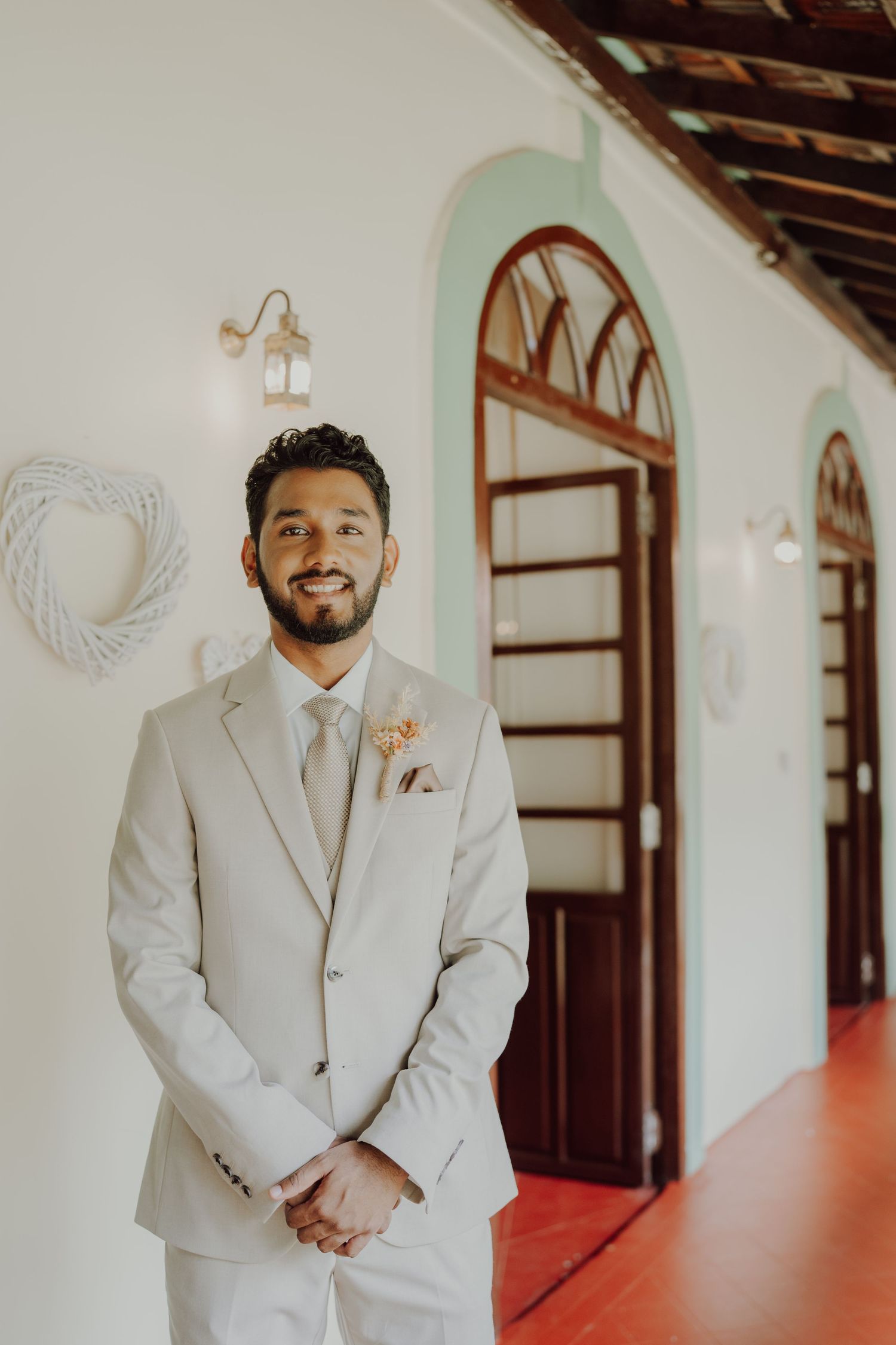 Smiling groom with a beard stands in a light beige suit with a floral boutonniere in a bright corridor with arched wooden doors.