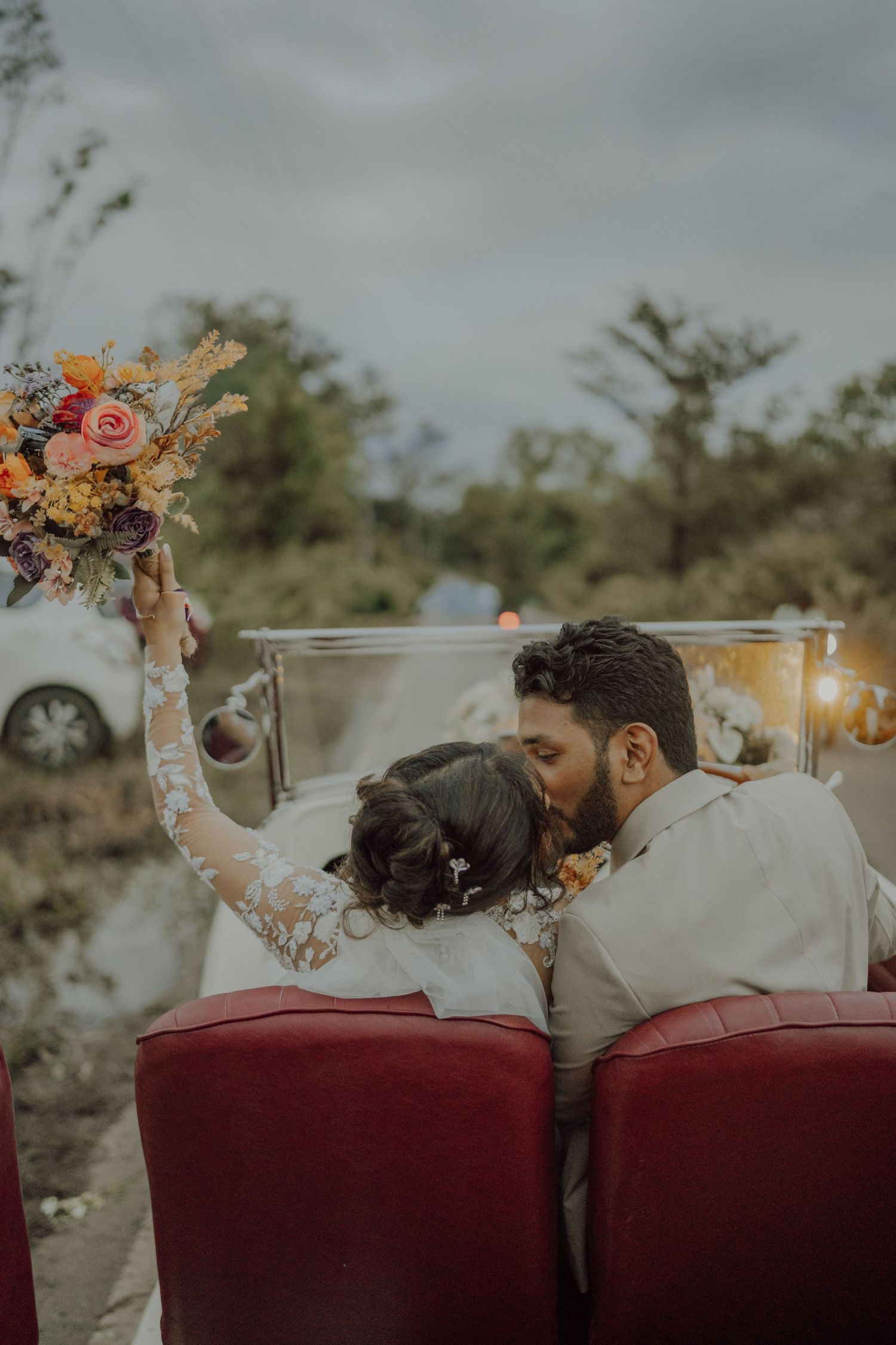 A groom kisses his bride from behind in a vintage convertible while she raises a colorful floral bouquet toward the cloudy sky.