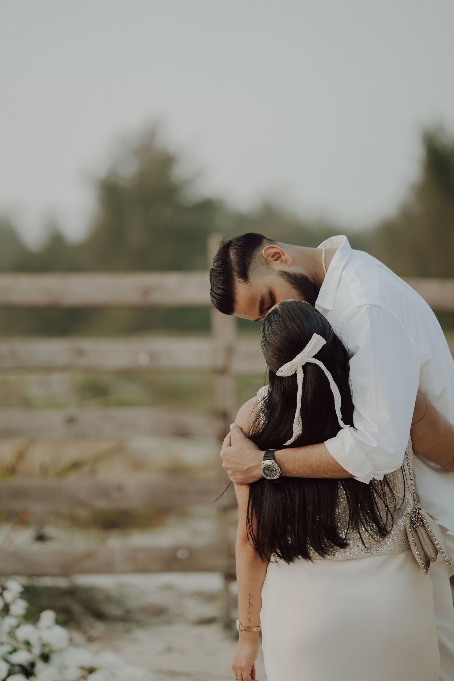 A couple embraces tenderly outdoors, the man in white kissing the woman's head near a rustic wooden fence.
