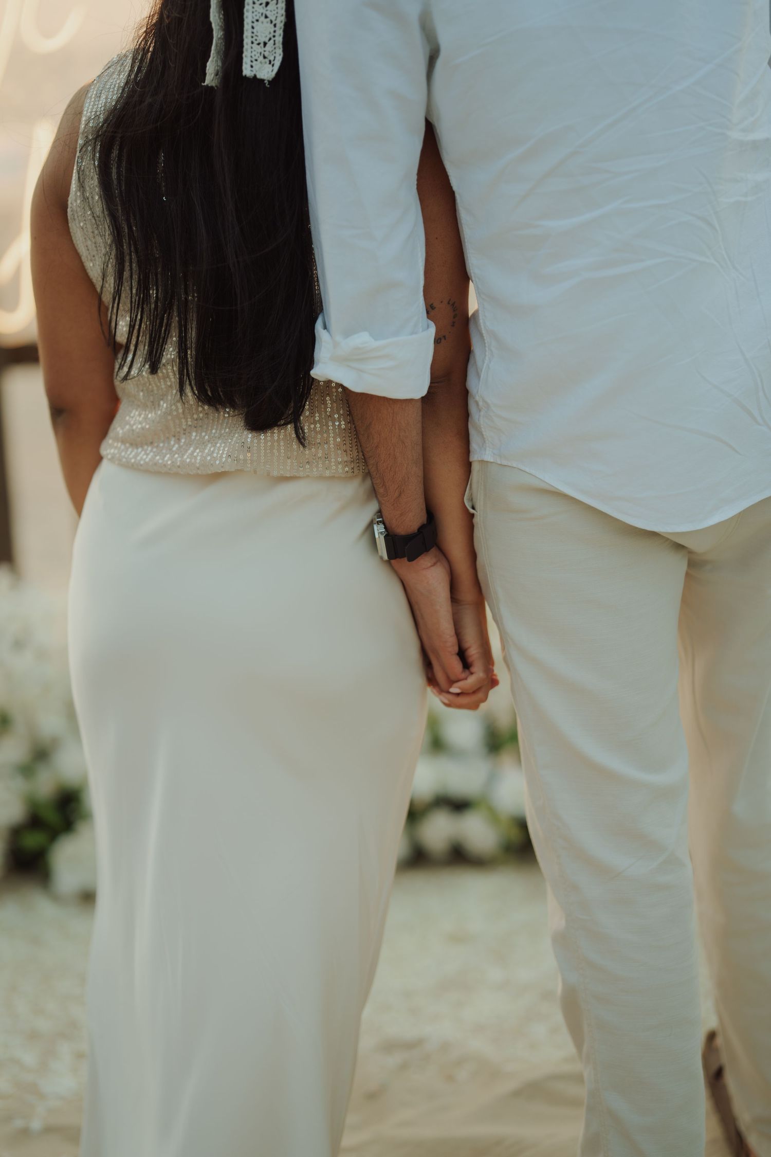 Couple holding hands from behind, wearing white outfits at a beach wedding ceremony.