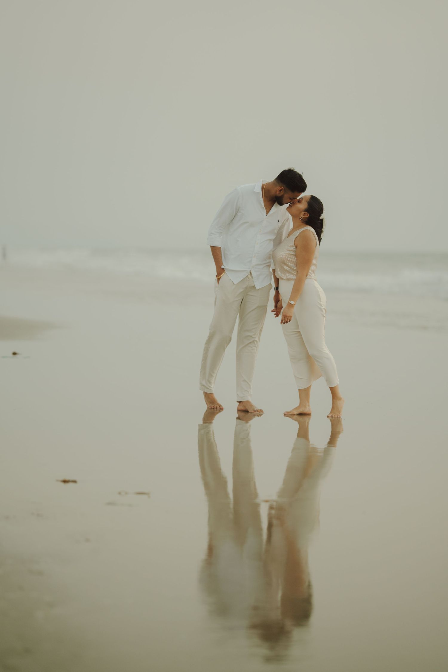 Couple in white outfits sharing a kiss on a reflective wet beach with ocean waves in the background.