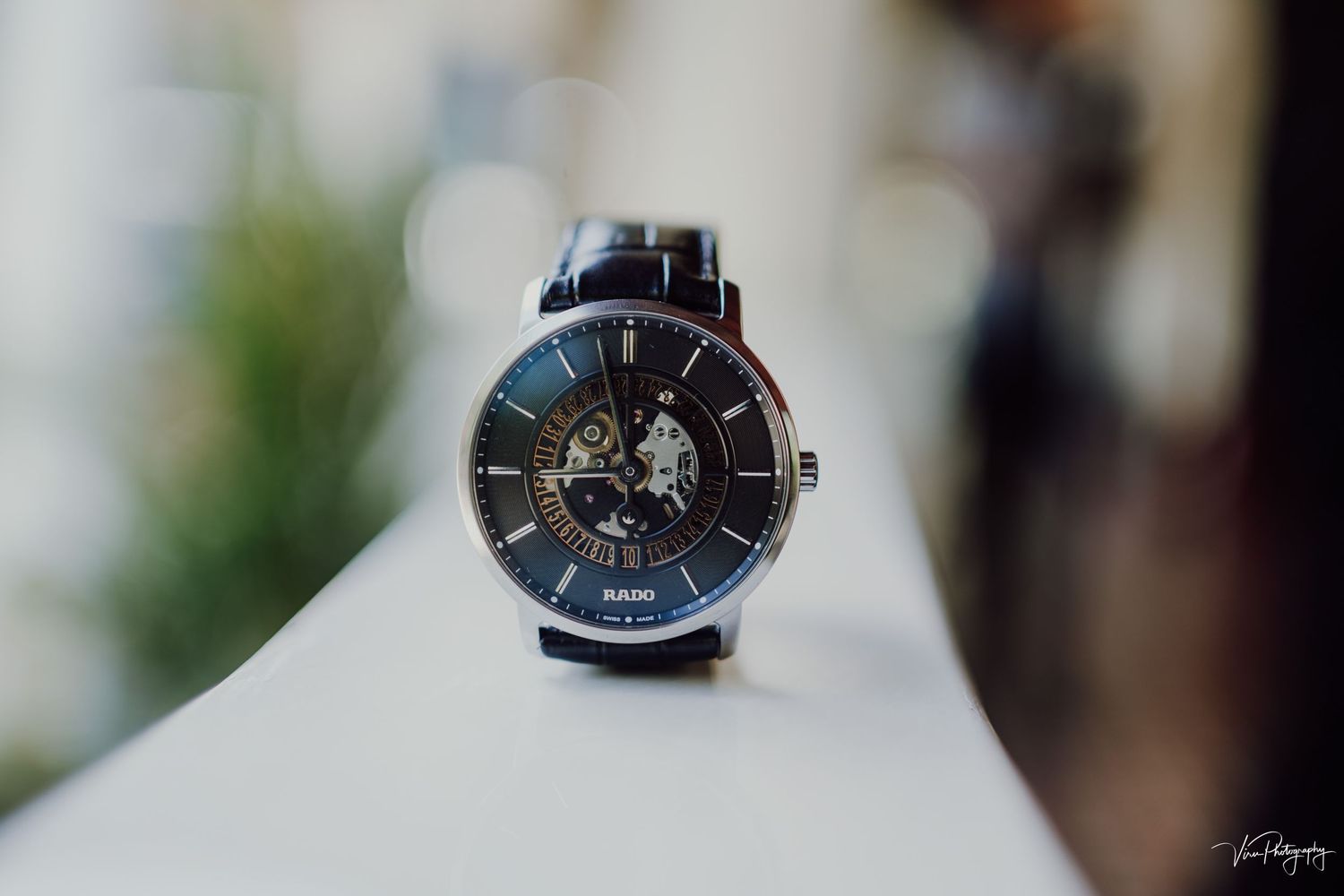 Close-up view of a luxury wristwatch with black leather strap and metallic skeleton dial face on white surface.