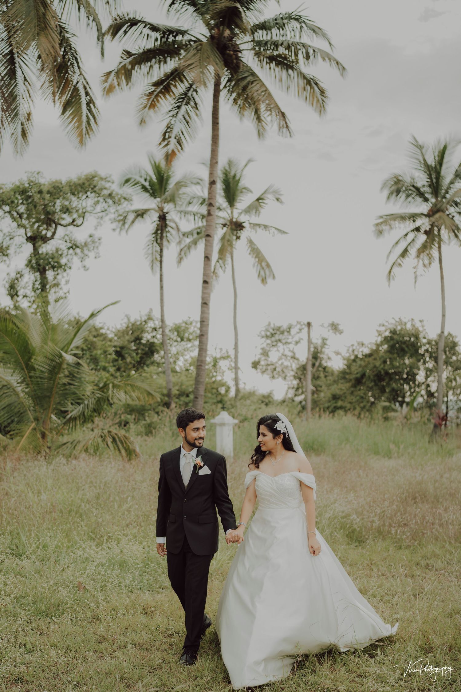 A couple walks together in a tropical garden setting with tall palm trees and lush greenery in the background.