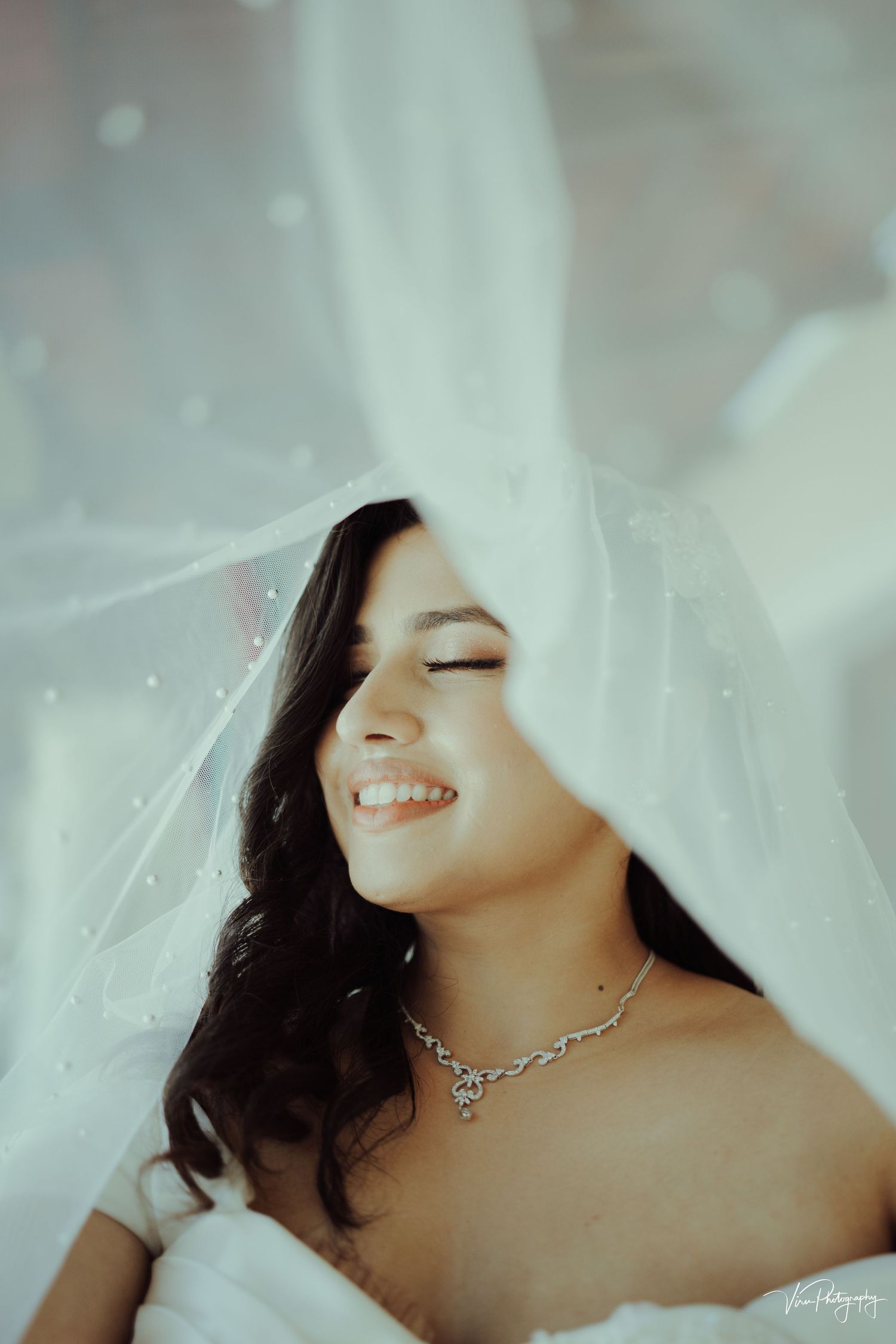 A joyful moment captured through delicate white wedding veil with sparkling necklace and soft lighting.