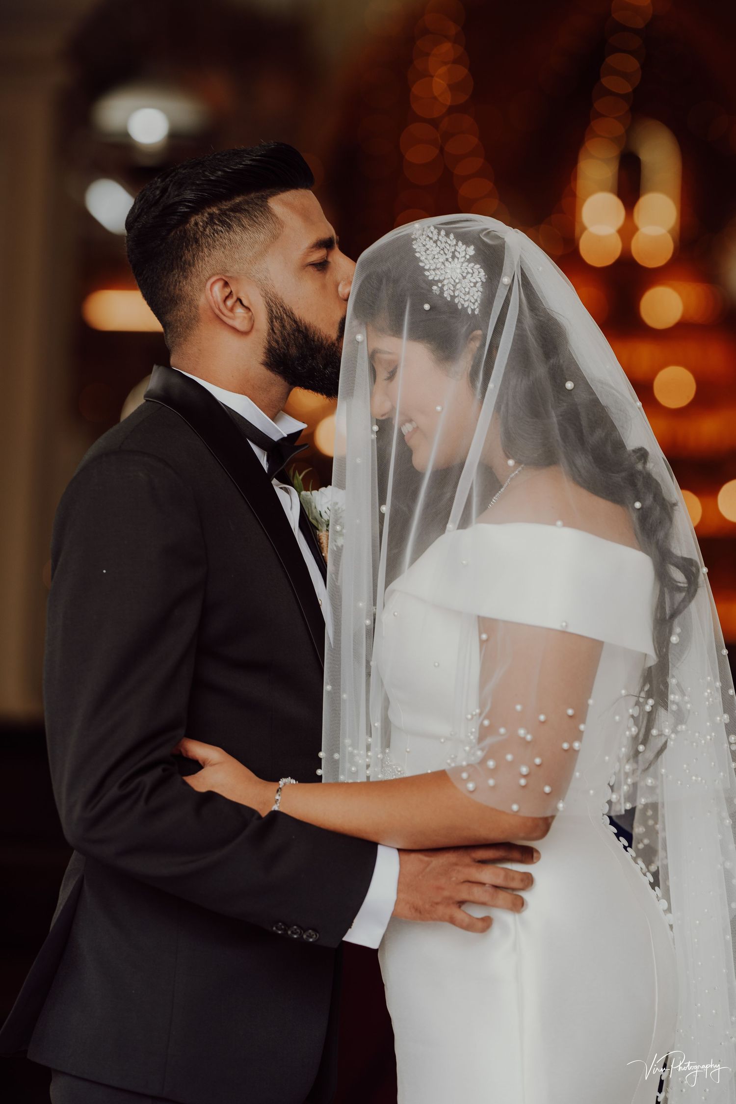 Romantic wedding moment captured with warm lighting as bride in elegant veil and groom share an intimate kiss.