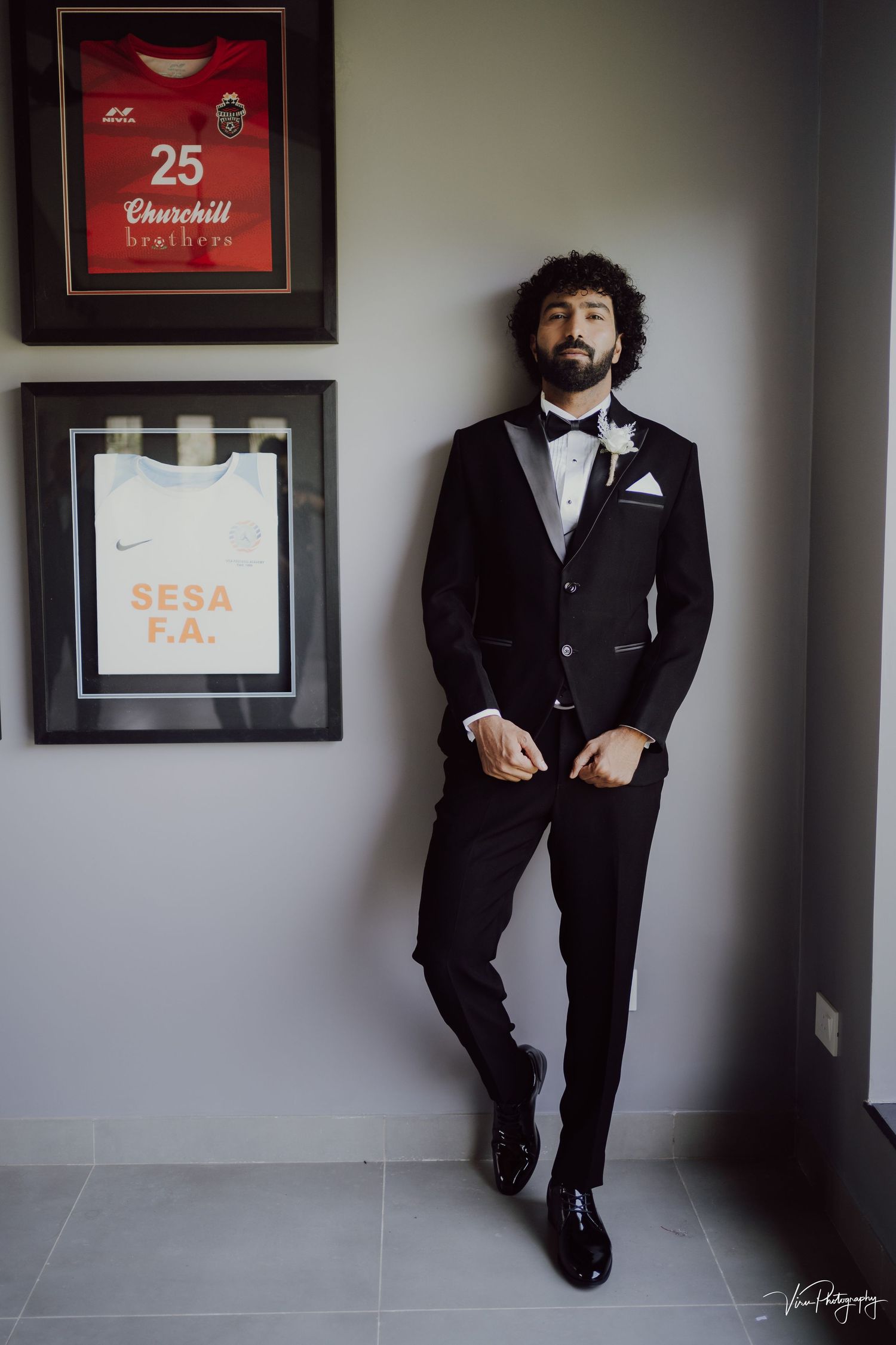 A person in a black tuxedo and bow tie poses against a gray wall with framed sports memorabilia.