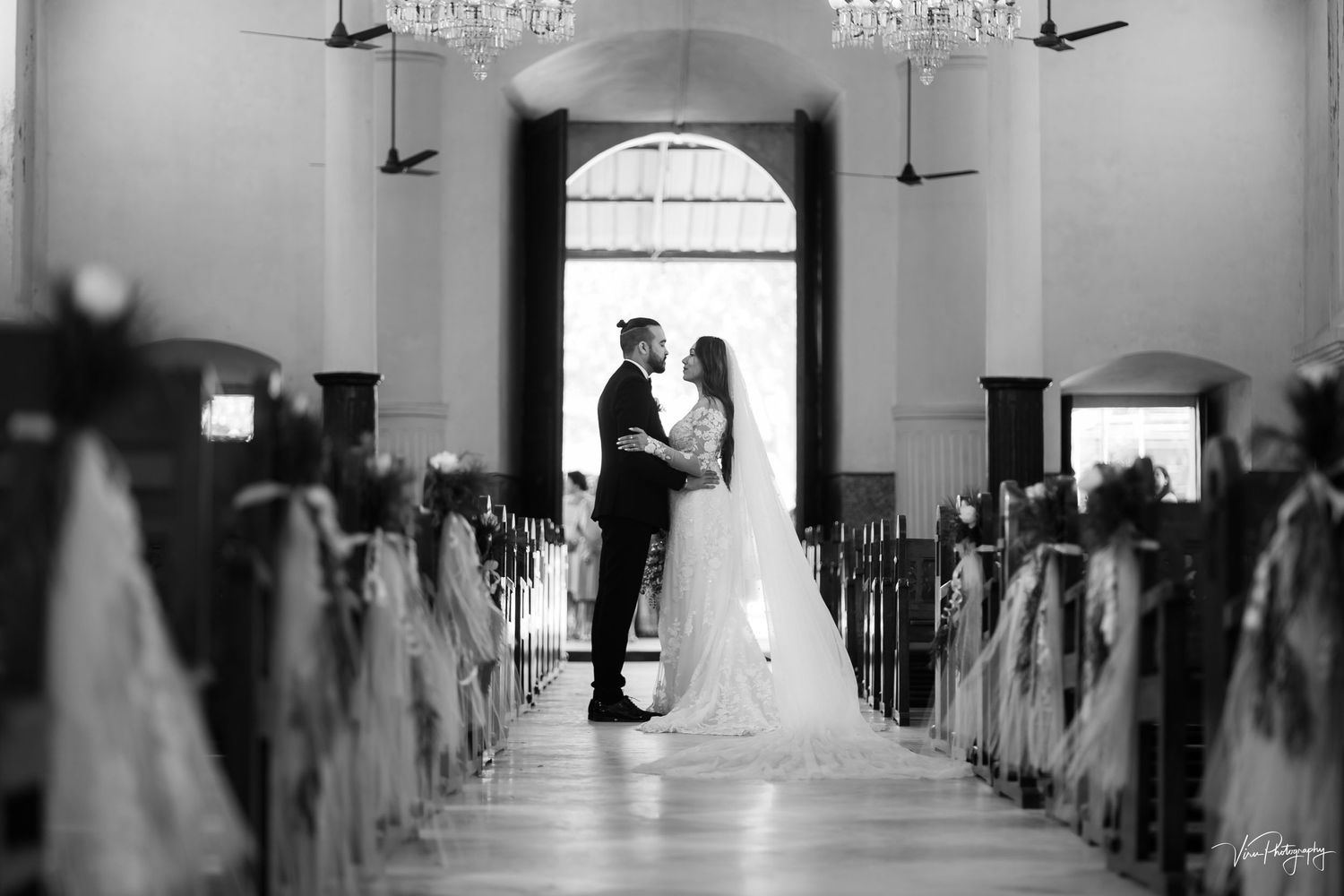 A black and white silhouette of a couple in wedding attire standing in a sunlit church aisle with decorated pews.