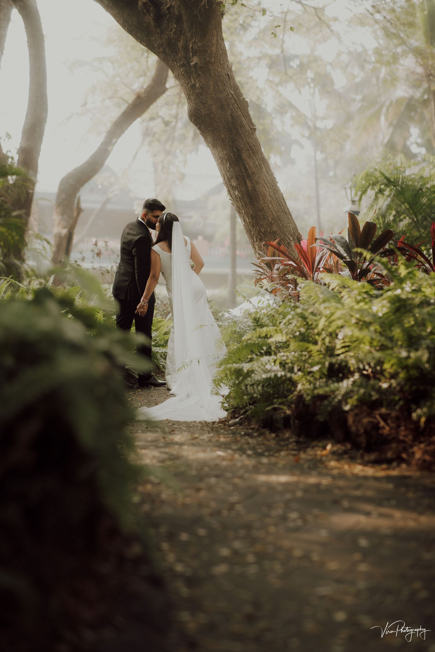 A romantic wedding couple embraces on a misty forest path surrounded by lush greenery and tropical plants.