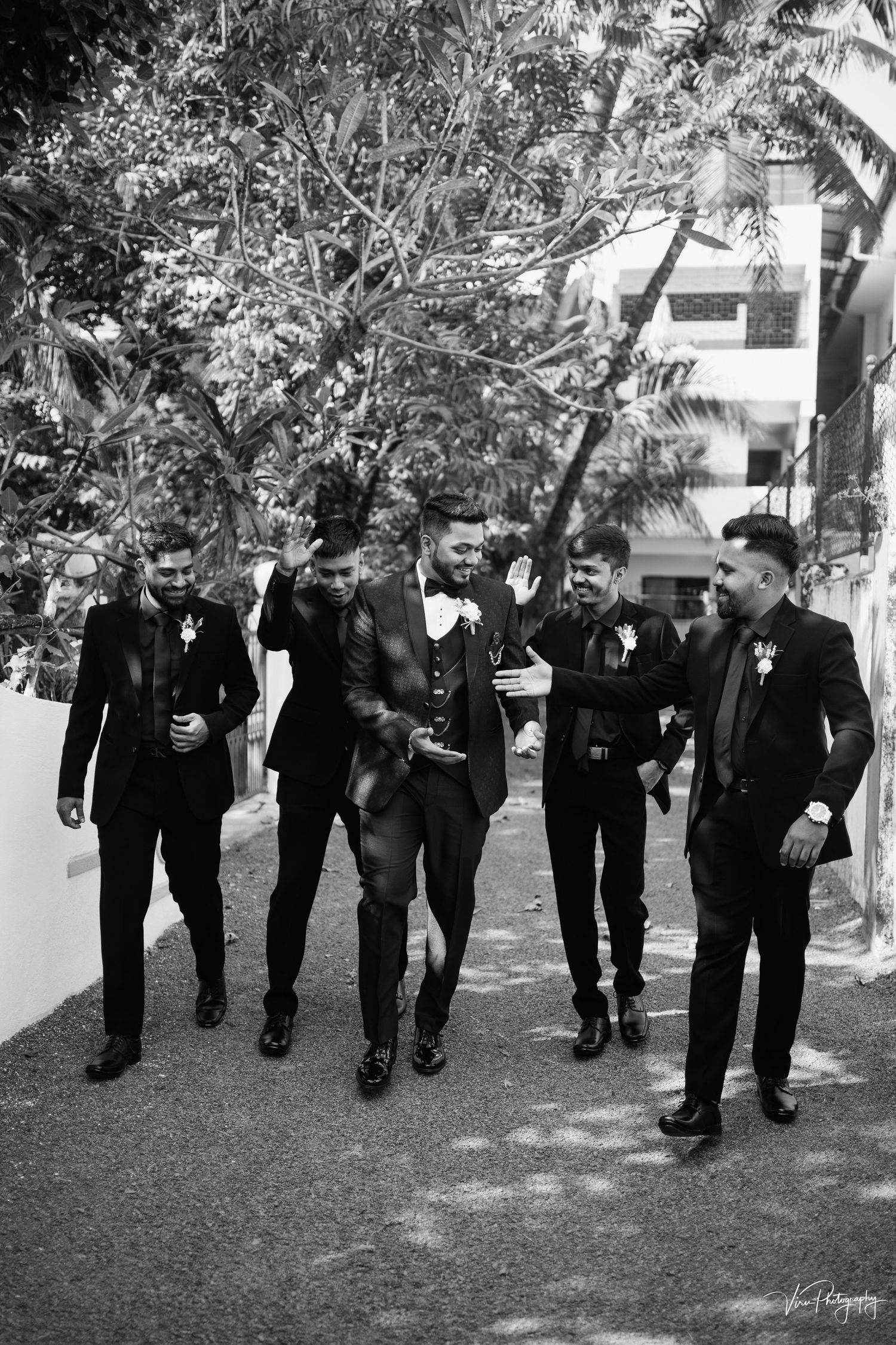 Five groomsmen in stylish black suits walking and smiling together on a tree-lined path outside a building.