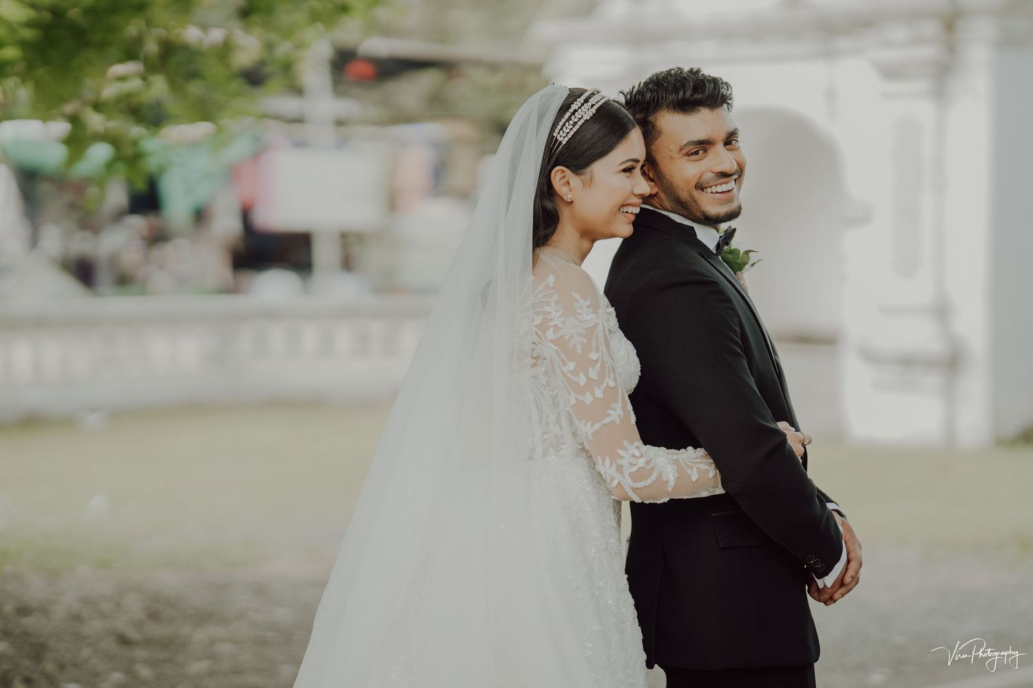 A bride in a white lace dress with long veil stands close to her groom in a black suit during an outdoor wedding.