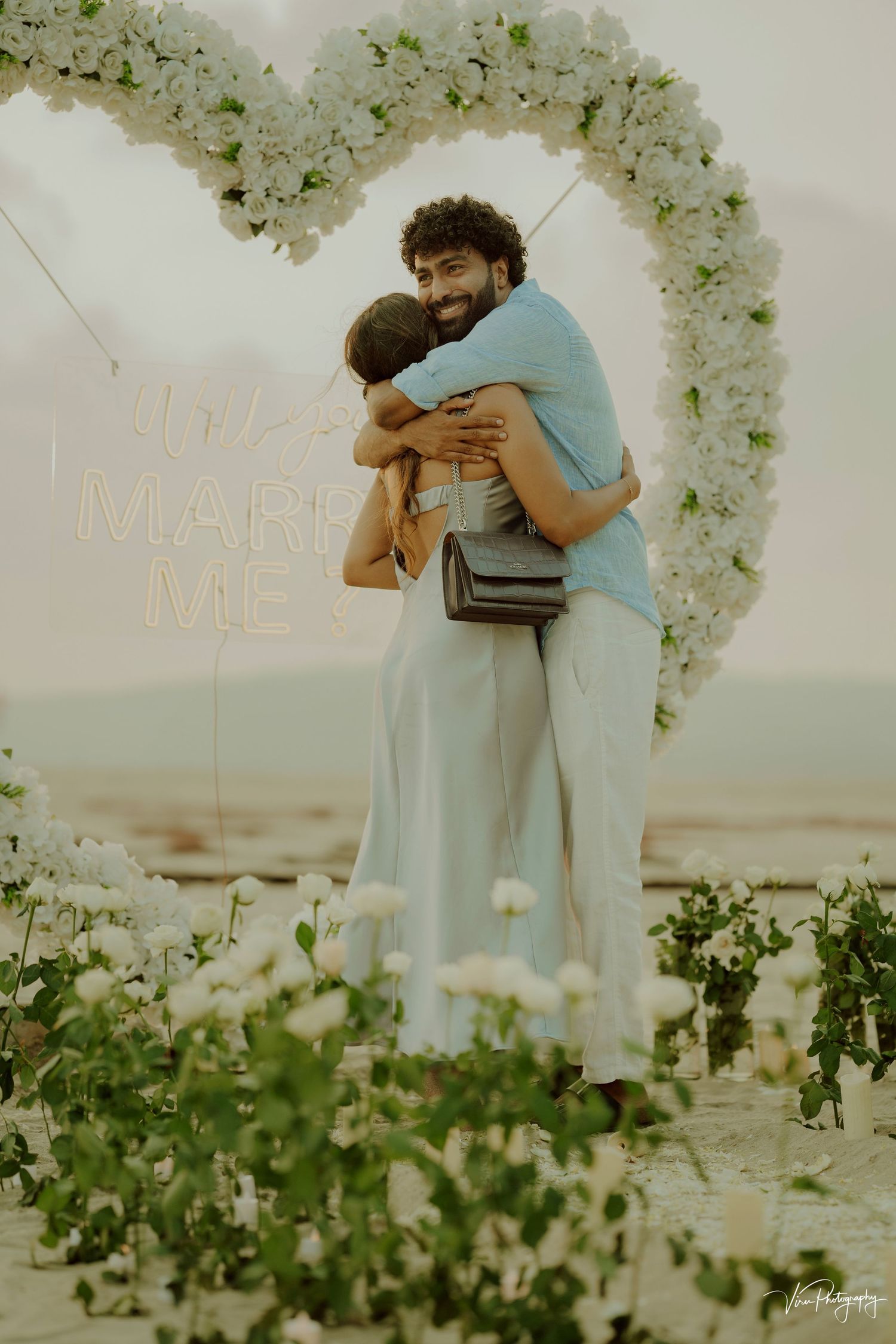 Two people embrace under a floral arch with 'Marry Me' text during a romantic beachside proposal at sunset.