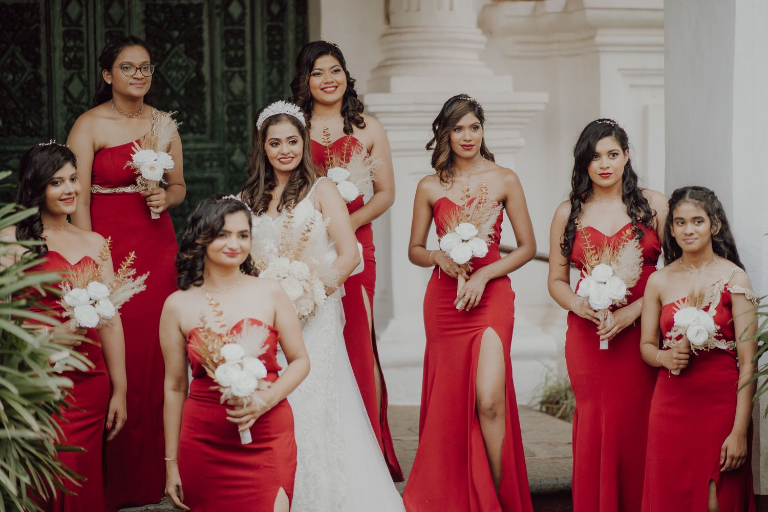 Wedding party in elegant red bridesmaid dresses and white bridal gown pose together with floral bouquets at venue.