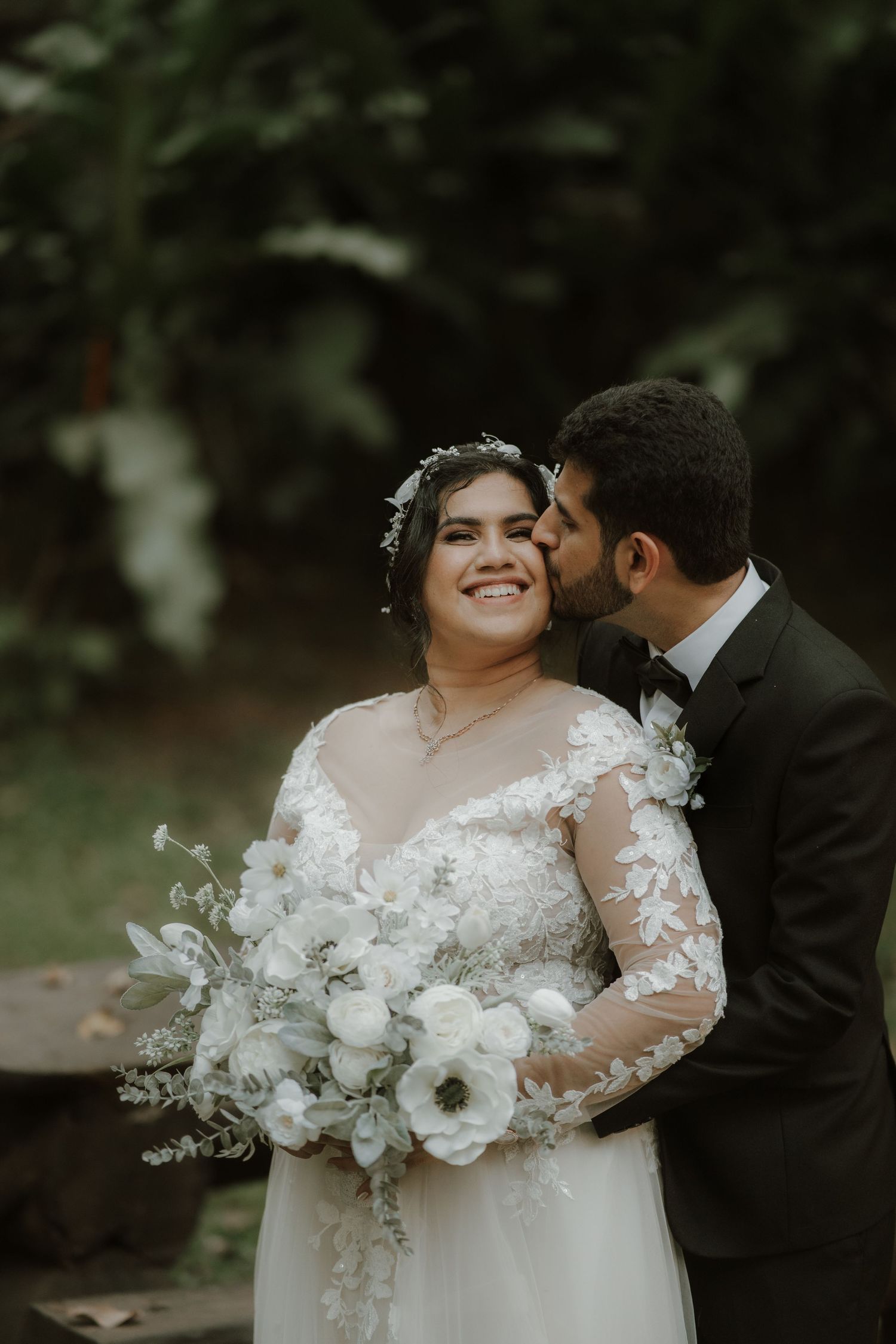 A romantic outdoor wedding photo of a couple in formal attire with the bride holding a white floral bouquet.