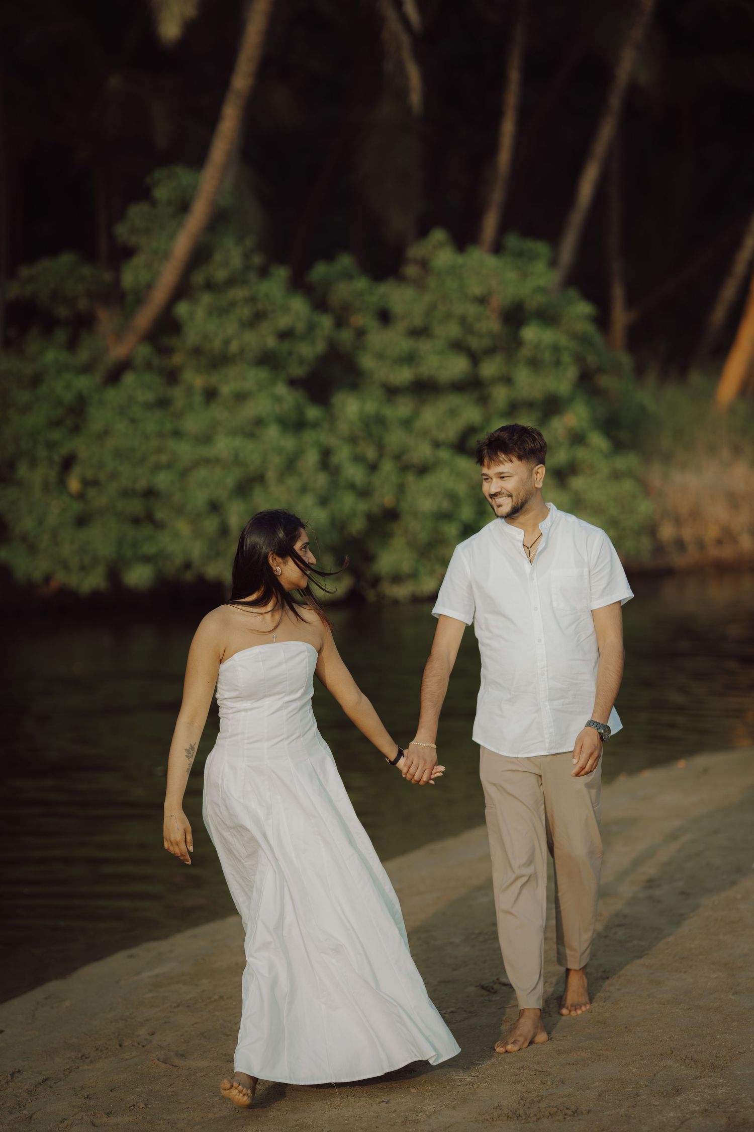 A couple in white and beige attire walks playfully along a beach at sunset with trees in the background.