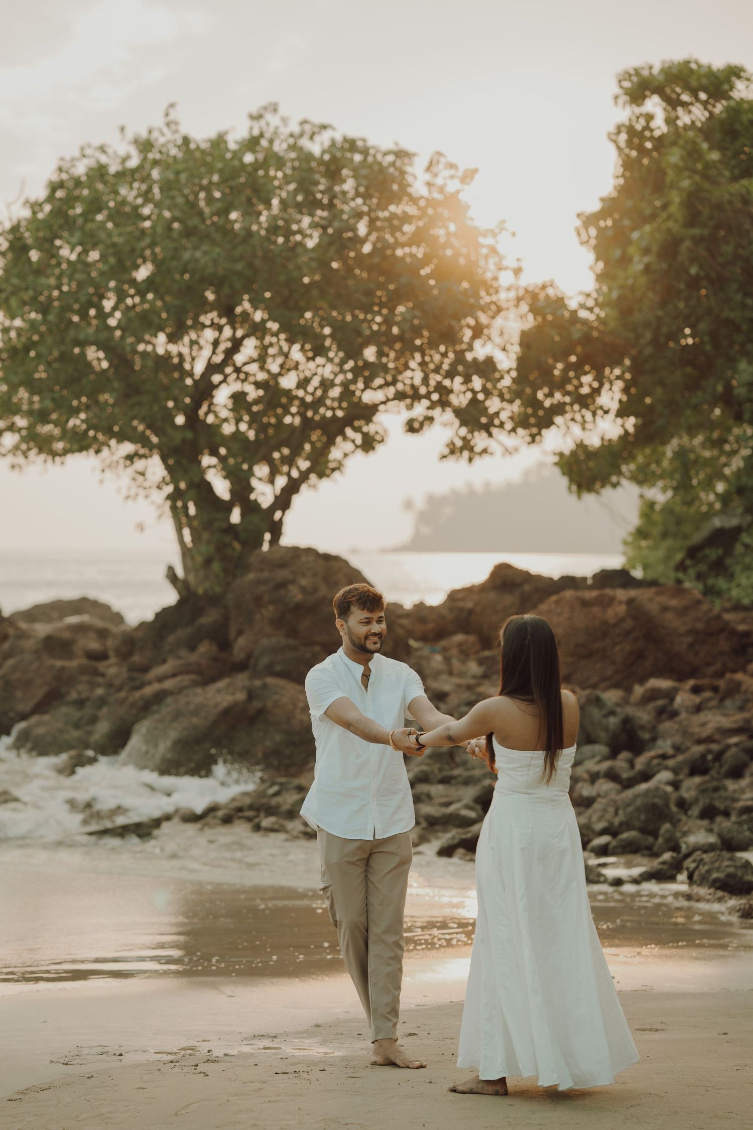 A couple walks along a rocky beach at sunset, with trees silhouetted against the golden sky.