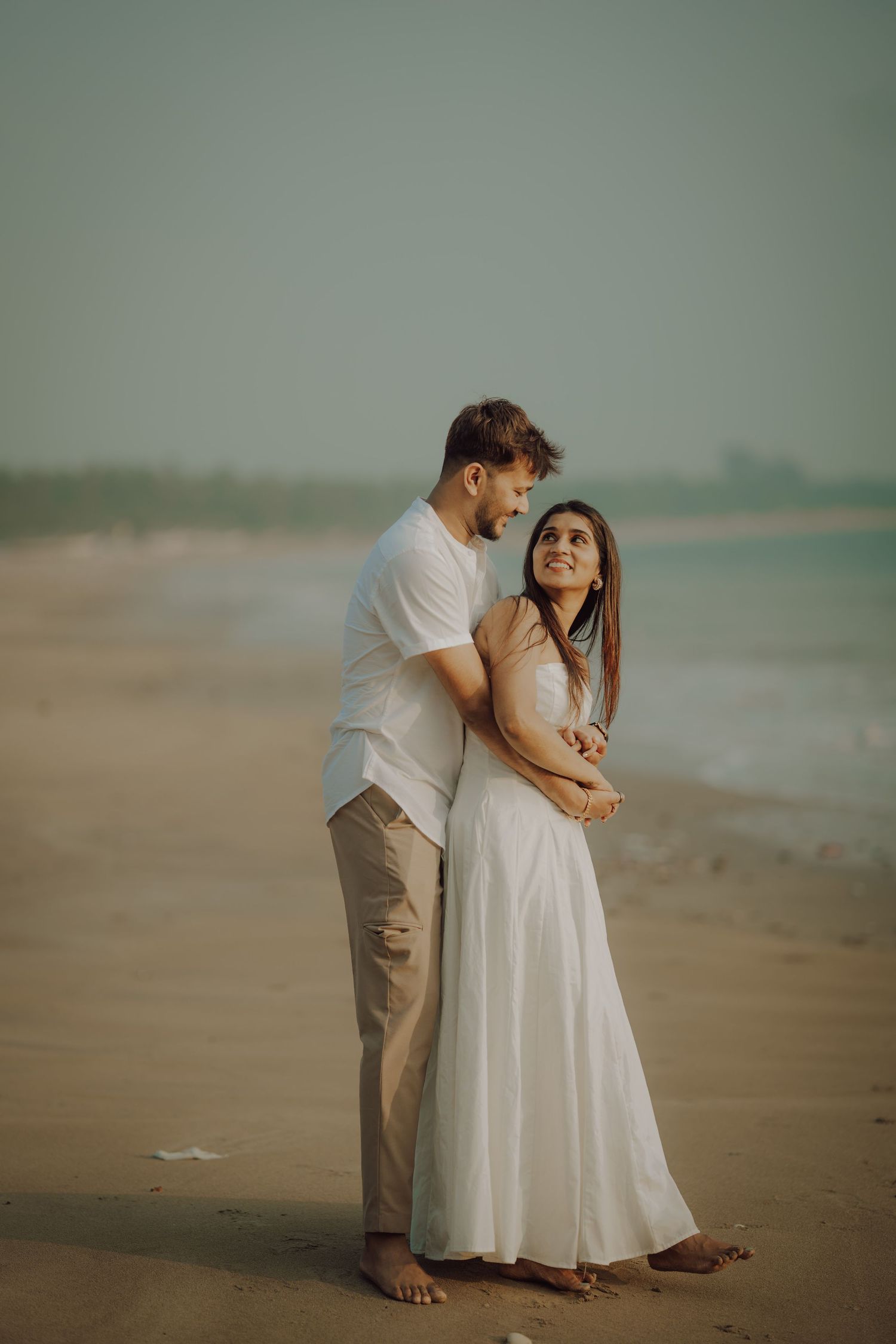 A couple shares a romantic moment on a misty beach, the woman wearing a flowing white dress and the man in casual attire.