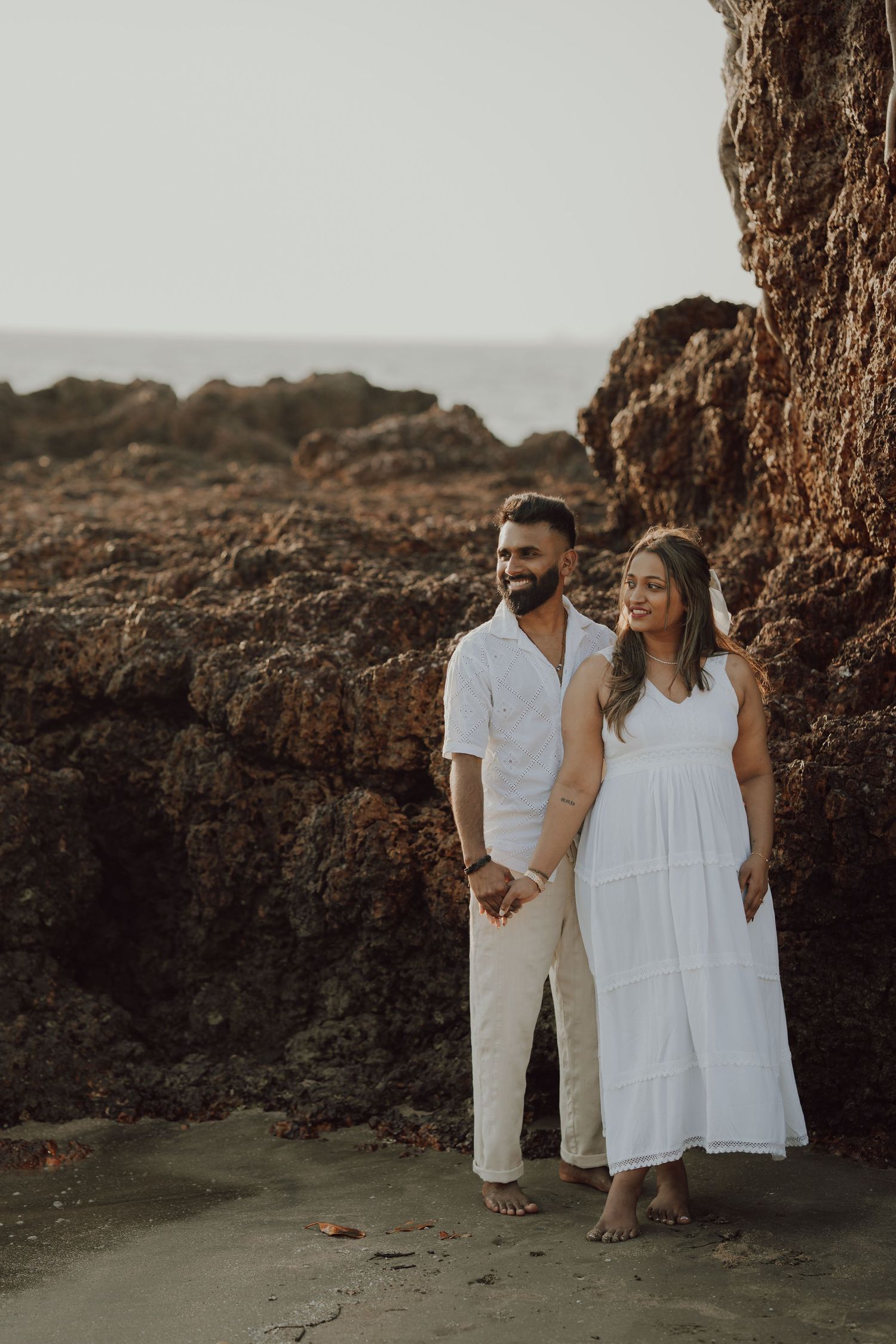 Couple in white outfits poses together against rocky coastal backdrop during a romantic photoshoot.