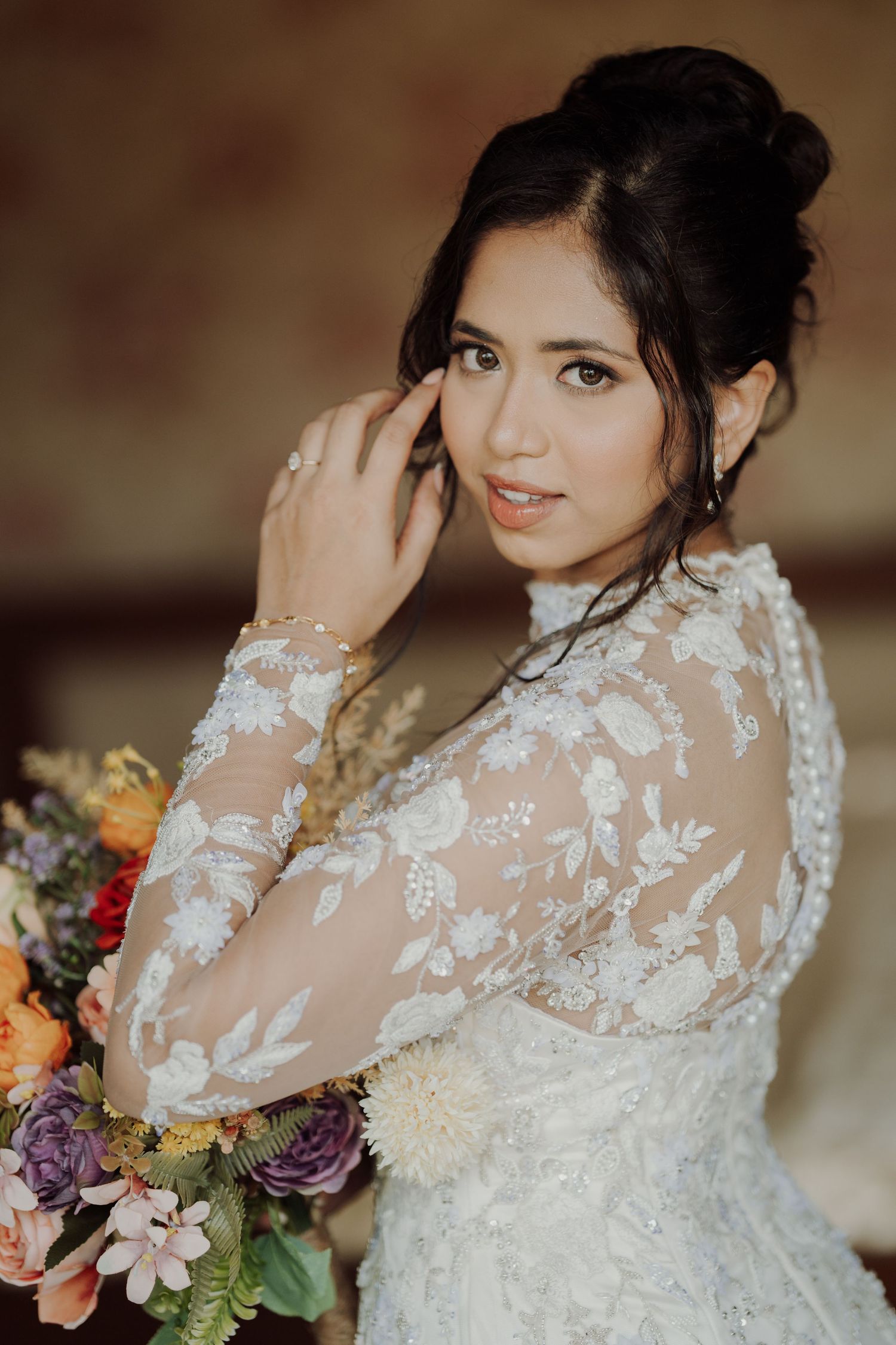 Bride in a long-sleeve white lace gown with an elegant updo posing beside a vibrant floral bouquet.