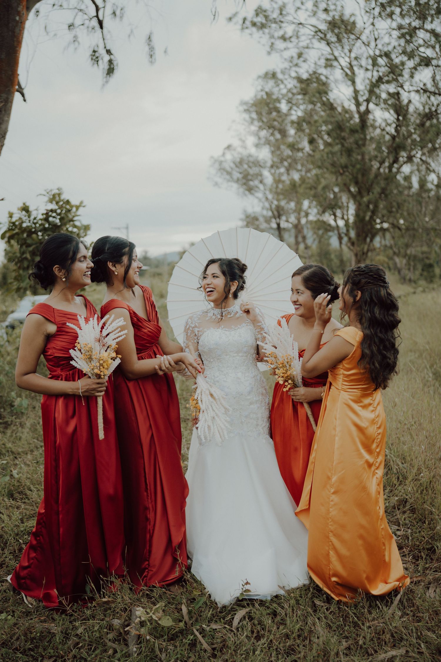 A bride in a white lace gown laughs with four bridesmaids in red and orange satin dresses holding dried flower bouquets outdoors.