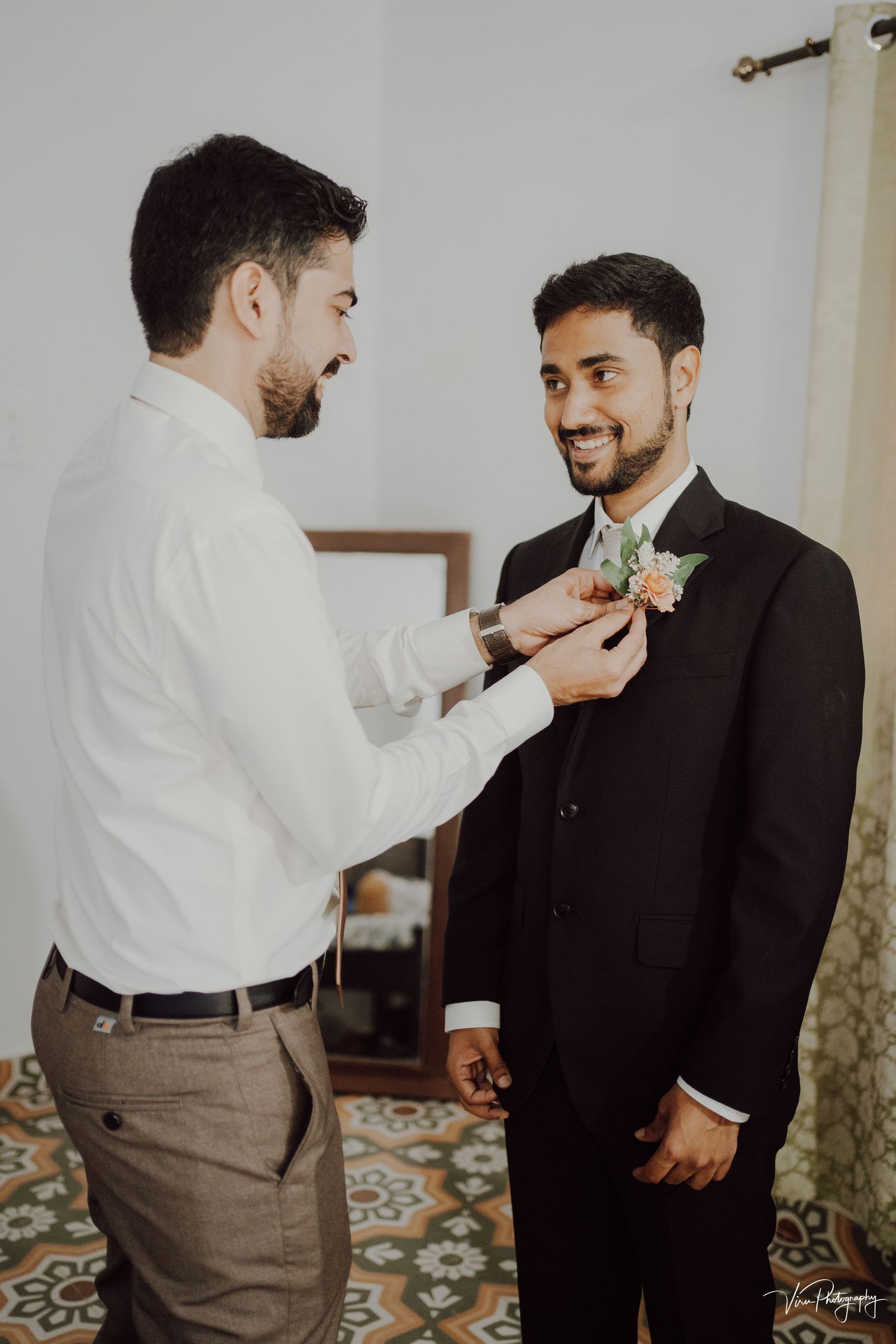 A groomsman helps adjust the boutonniere on a smiling groom's black suit lapel during wedding preparations.