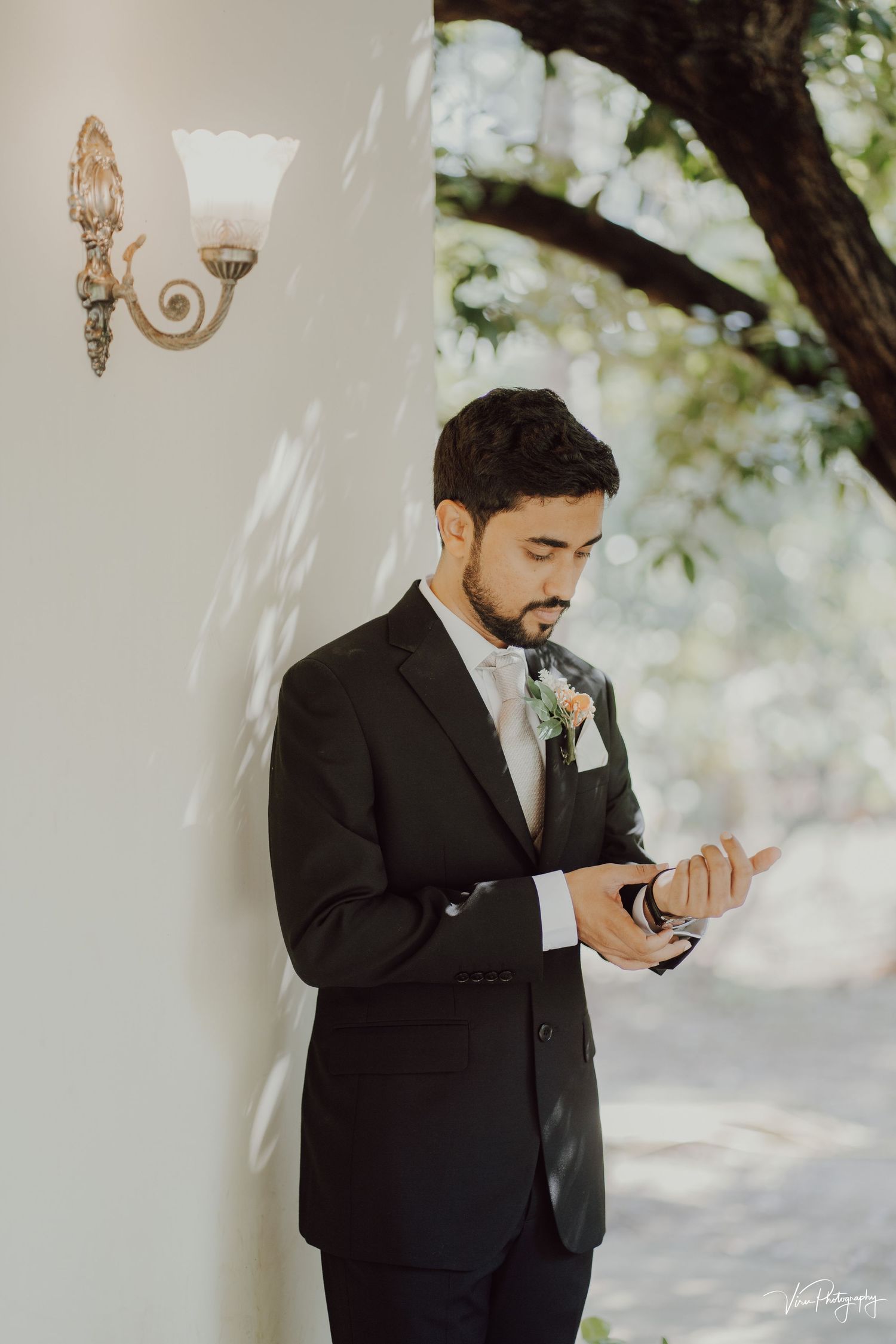 A groom in a classic black tuxedo checks his phone while standing against a white wall with an ornate sconce.