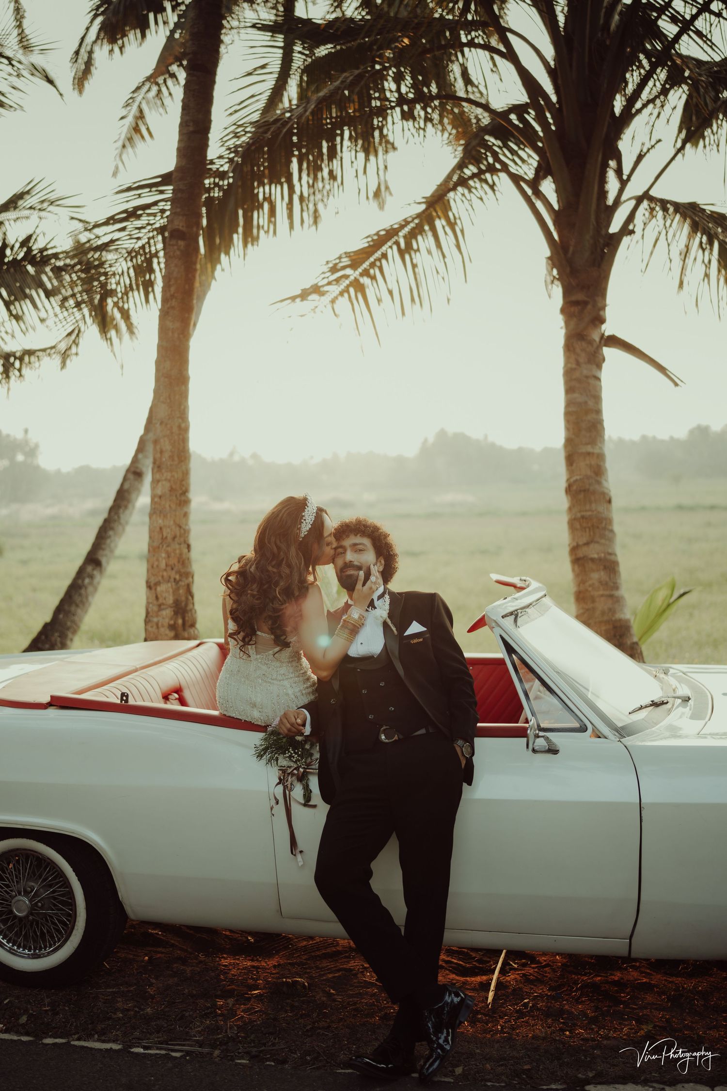 A romantic wedding couple poses with a classic white convertible car under palm trees at sunset.