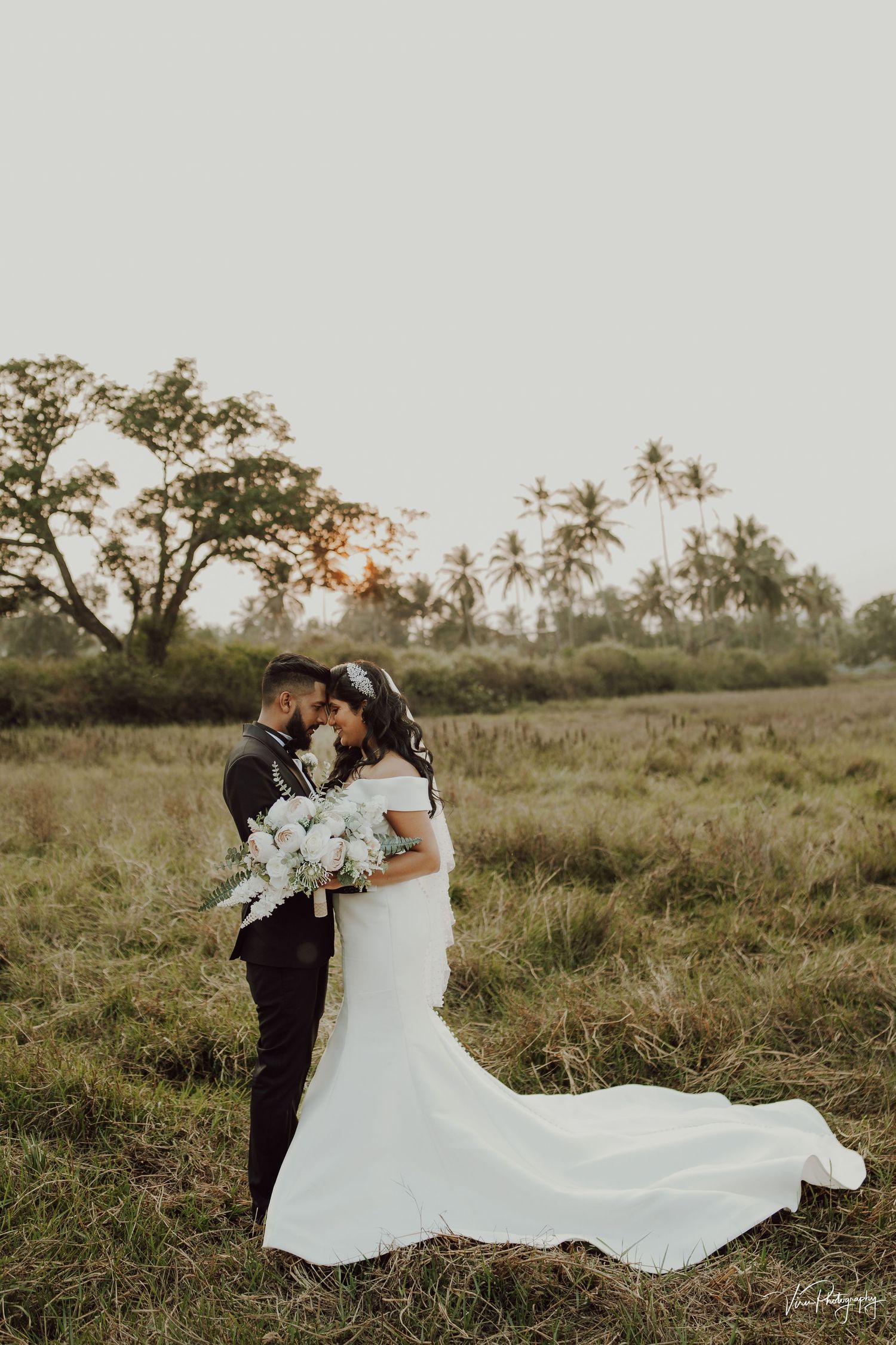 A couple embraces in a romantic sunset field with palm trees and flowing white wedding dress trailing behind them.