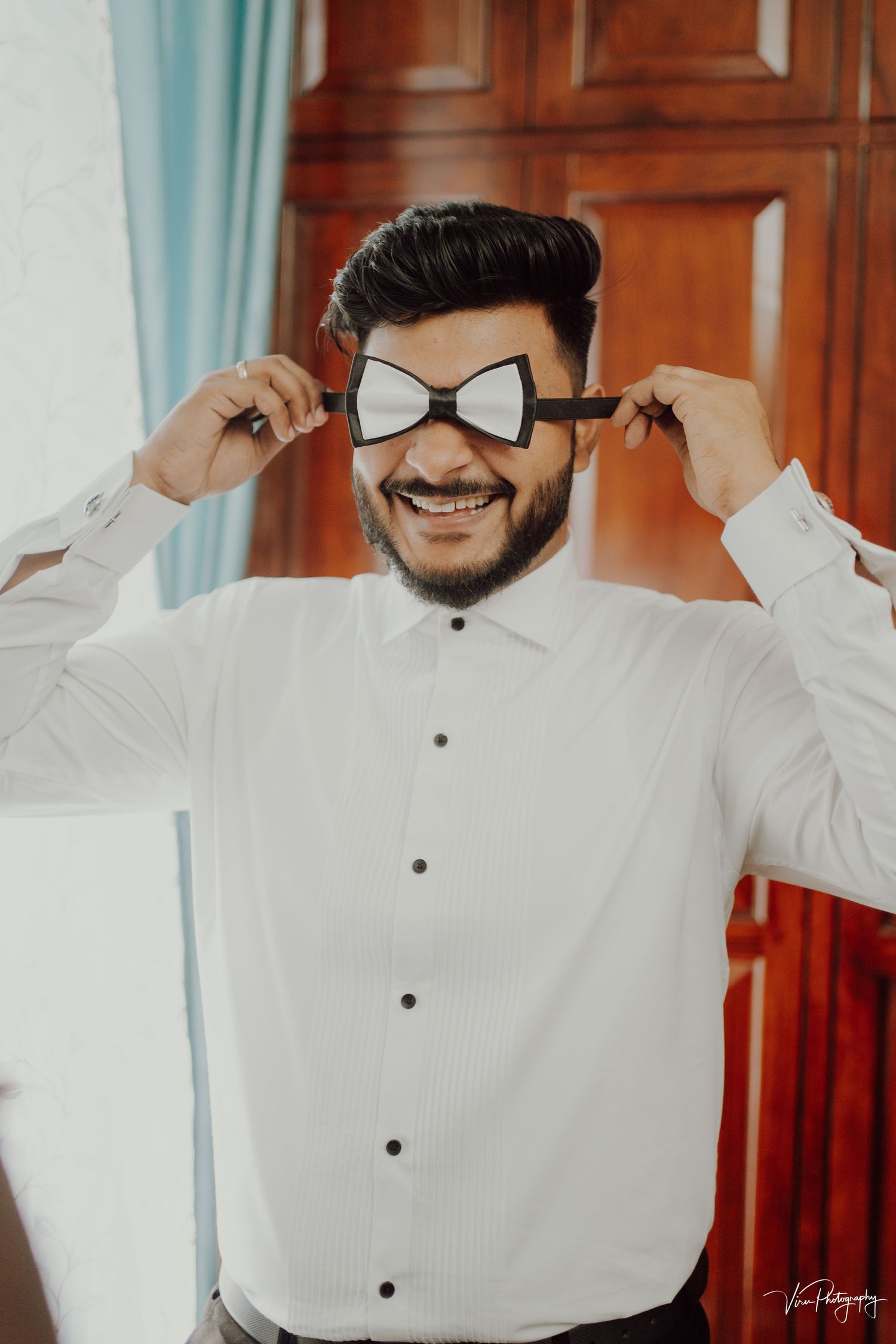 Someone adjusts their classic black and white bowtie while wearing a crisp white dress shirt and smiling warmly.