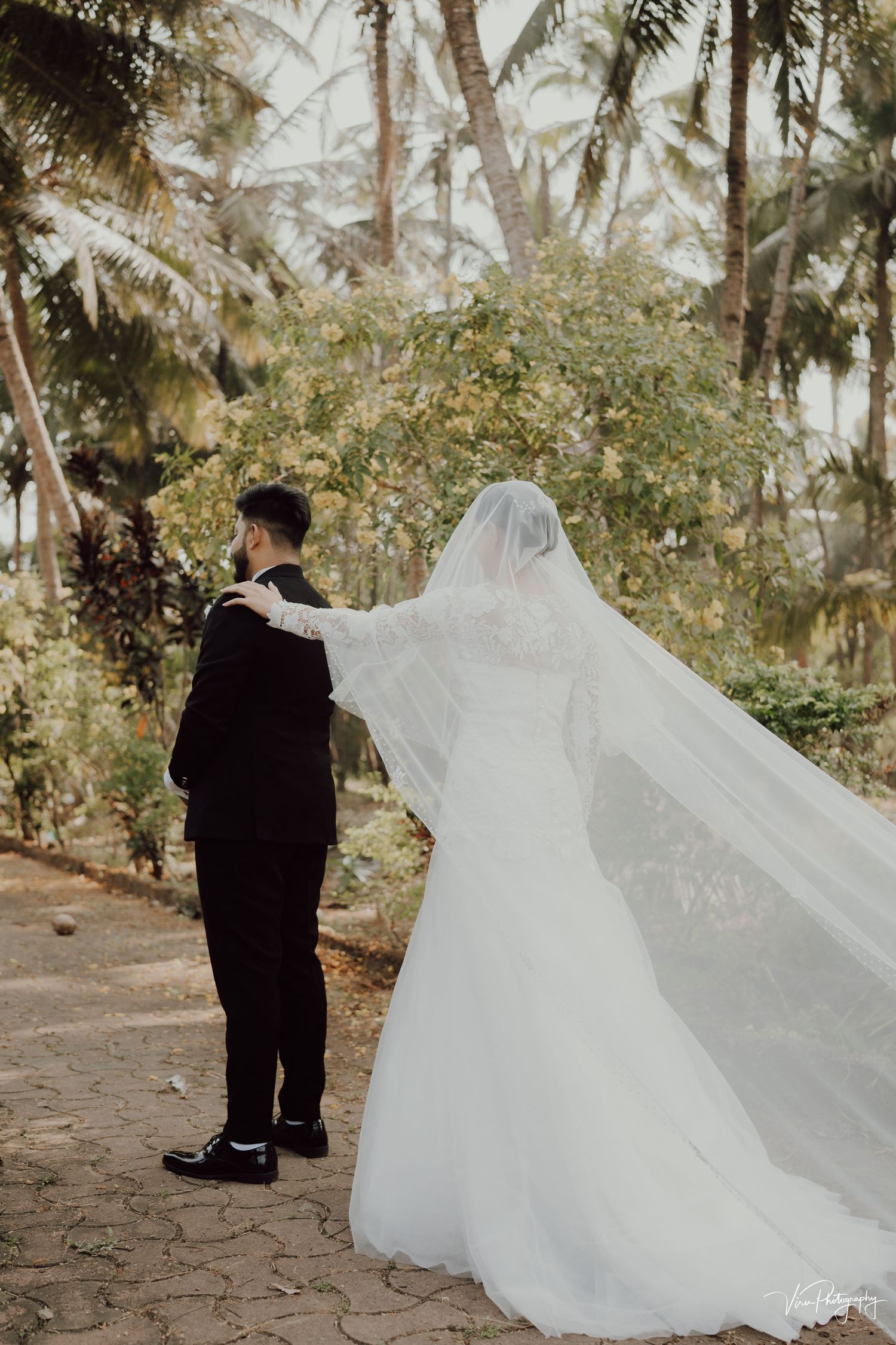 A bride in flowing white gown and veil stands with groom among tropical palm trees on a stone path.