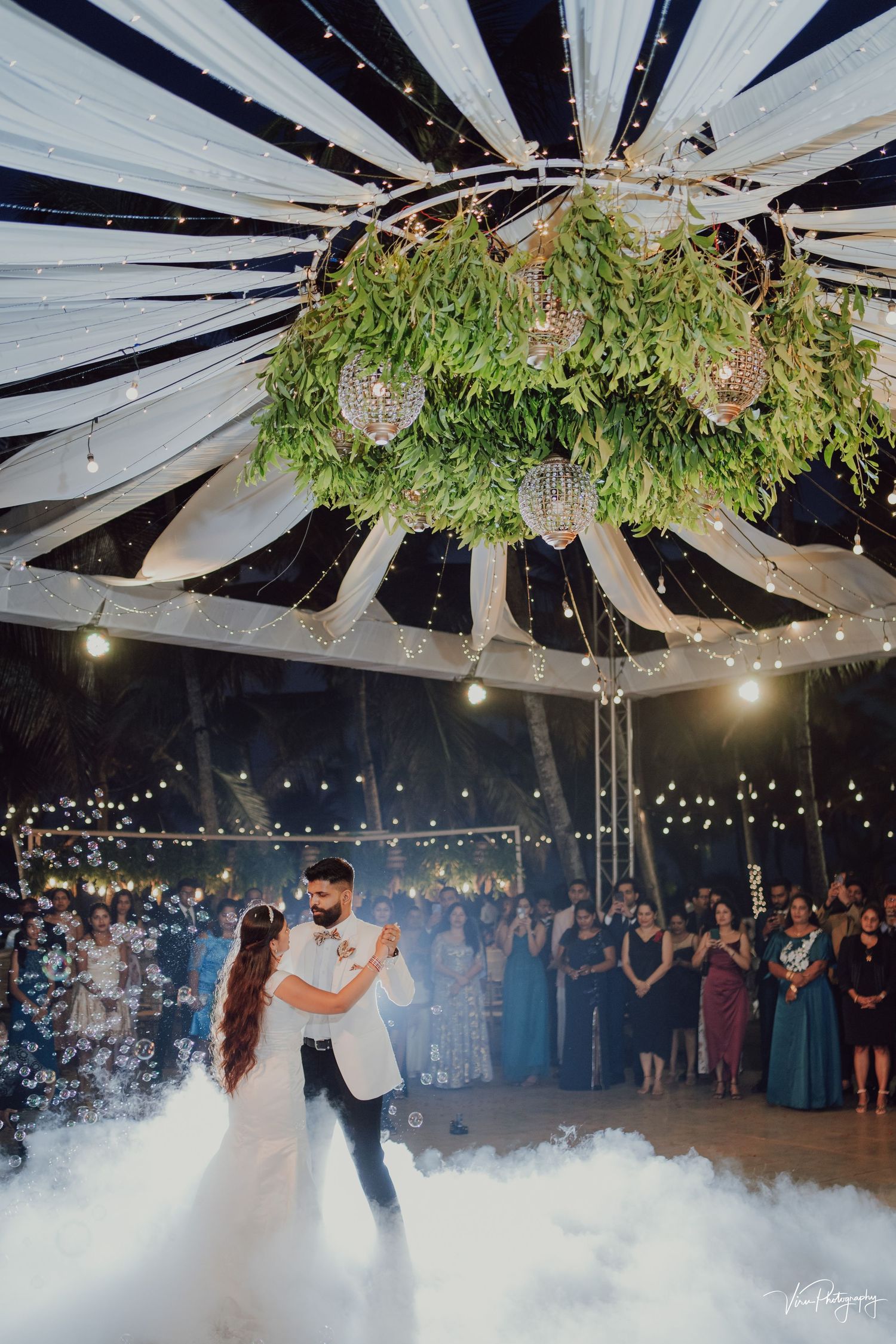 A couple shares their first dance under a moss and floral chandelier with dramatic fog effects and twinkling lights at a wedding reception.