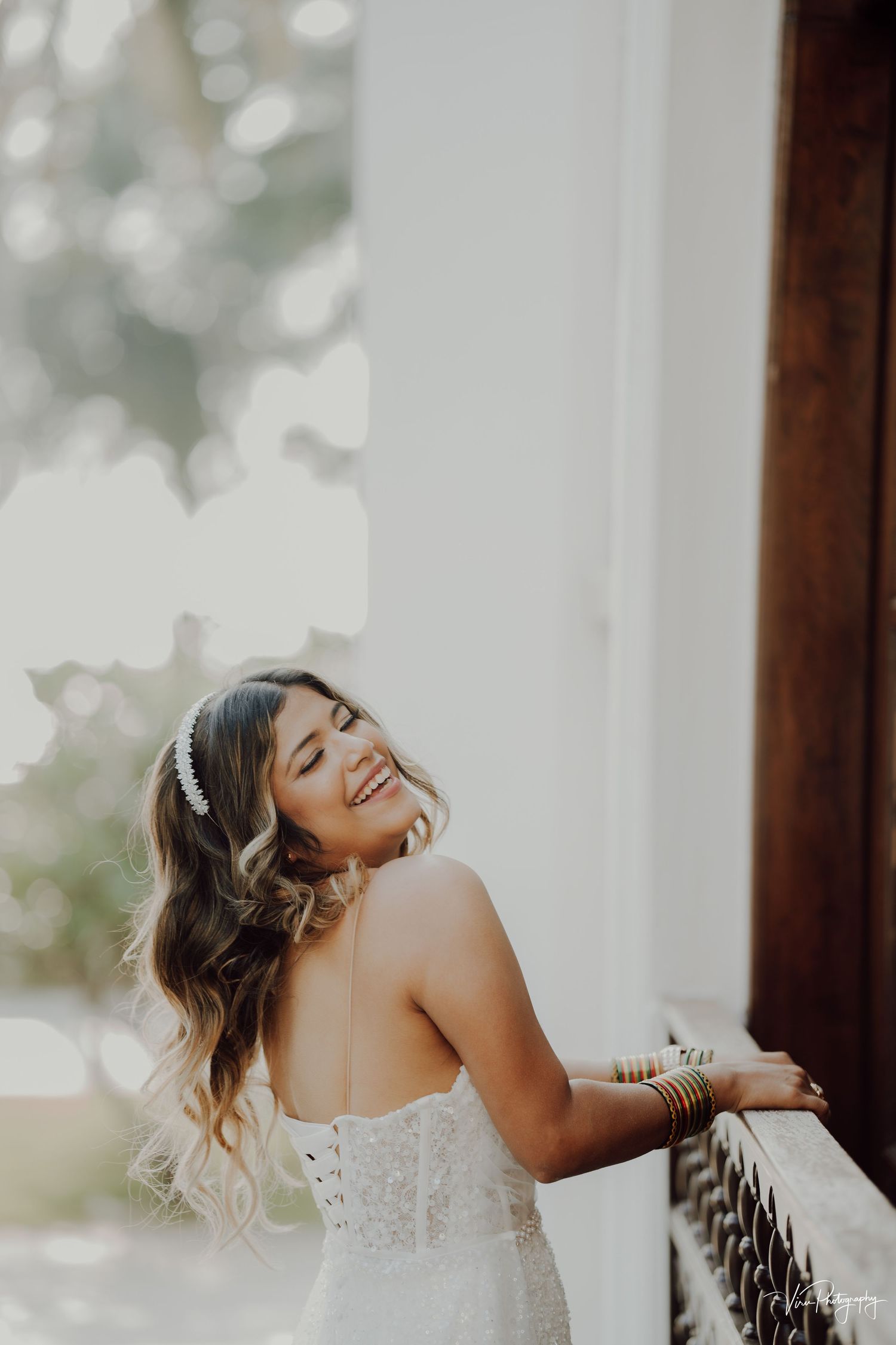 A bride in a white lace wedding dress laughs joyfully while standing next to an ornate wooden railing in soft light.