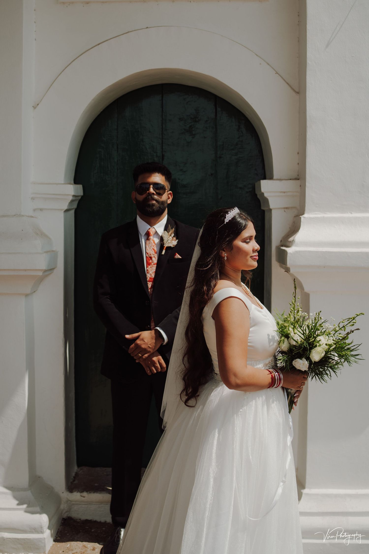 A bride in a white gown holds a bouquet while standing near an arched doorway as part of a wedding ceremony.