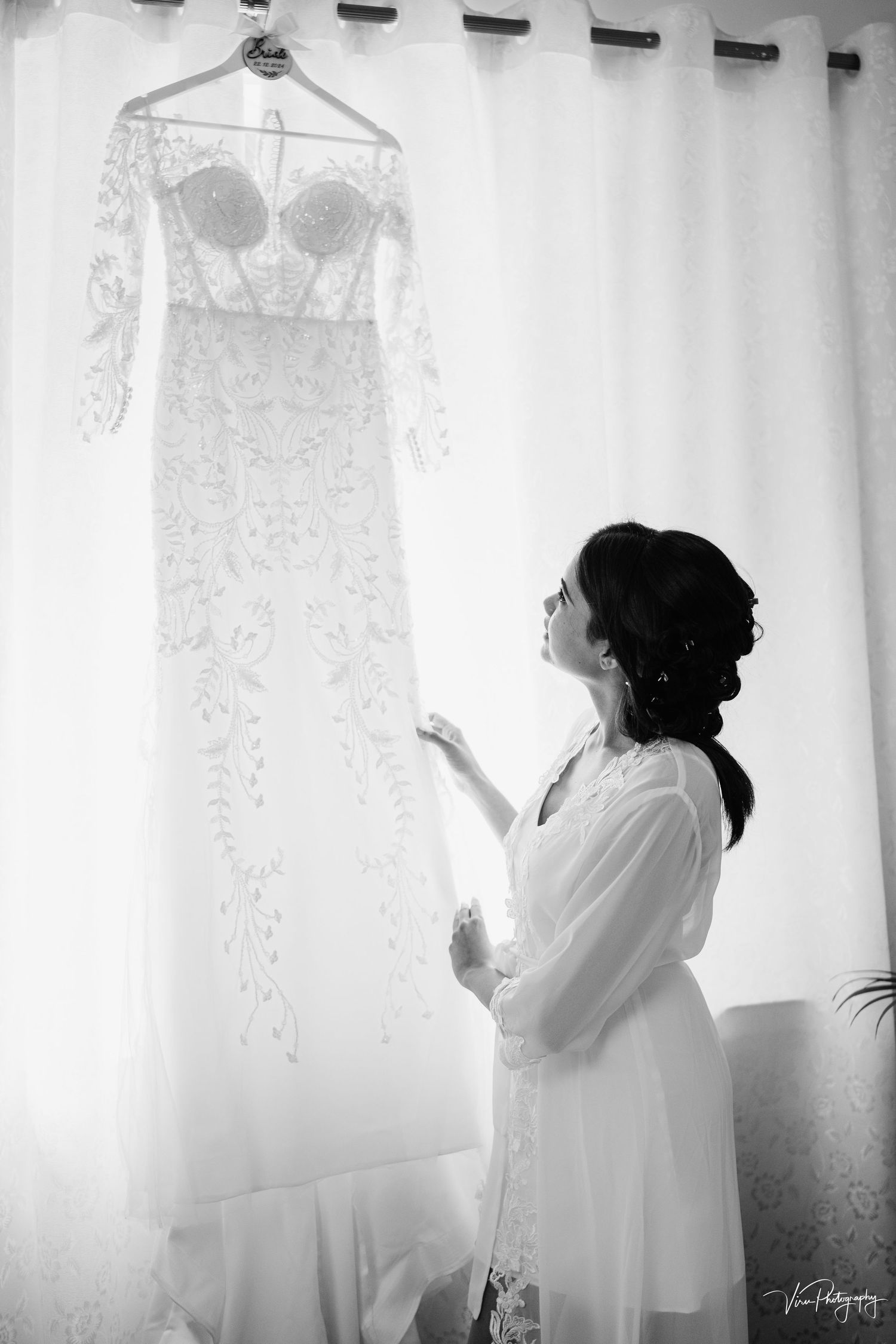 A black and white photo showing a delicate lace wedding dress on a hanger while someone admires it against sheer curtains.