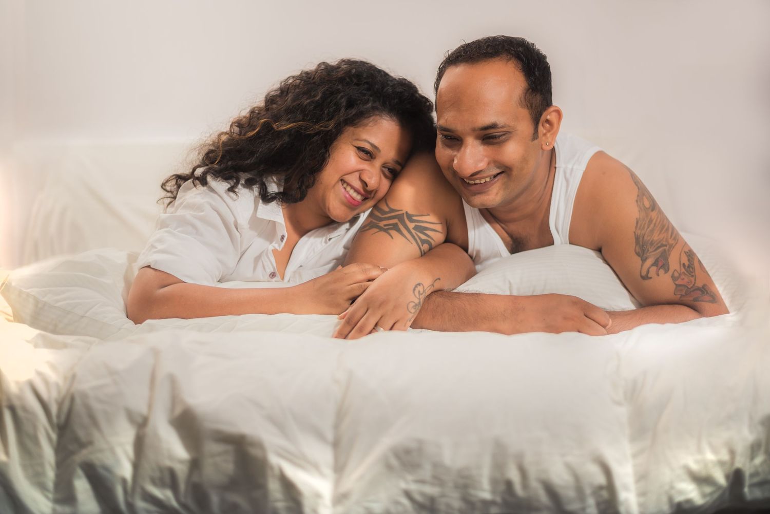 Couple lying together on white bedding while sharing an intimate moment and laughing.