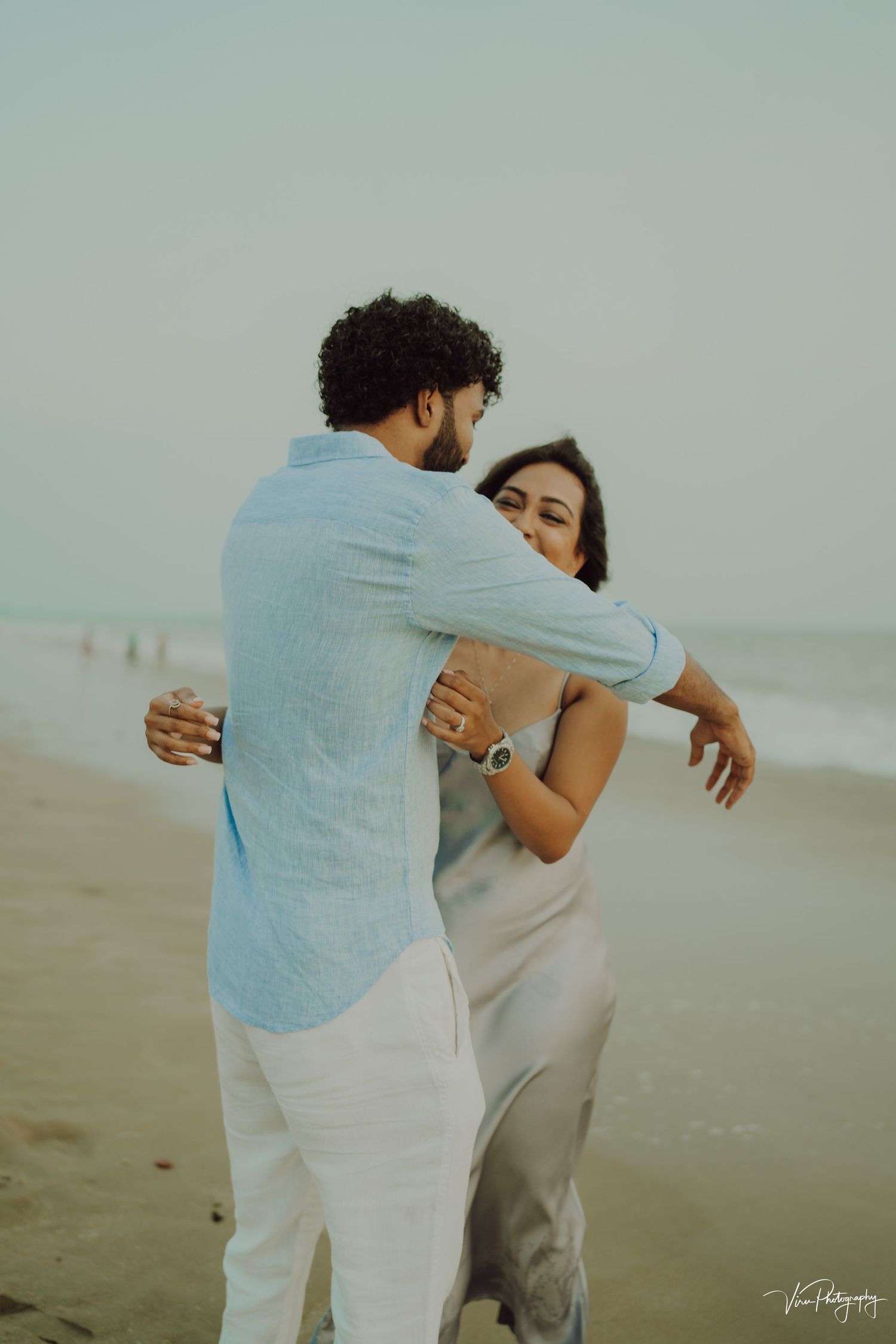 Romantic couple embraces on misty beach at sunset wearing light colored outfits in warm tones.