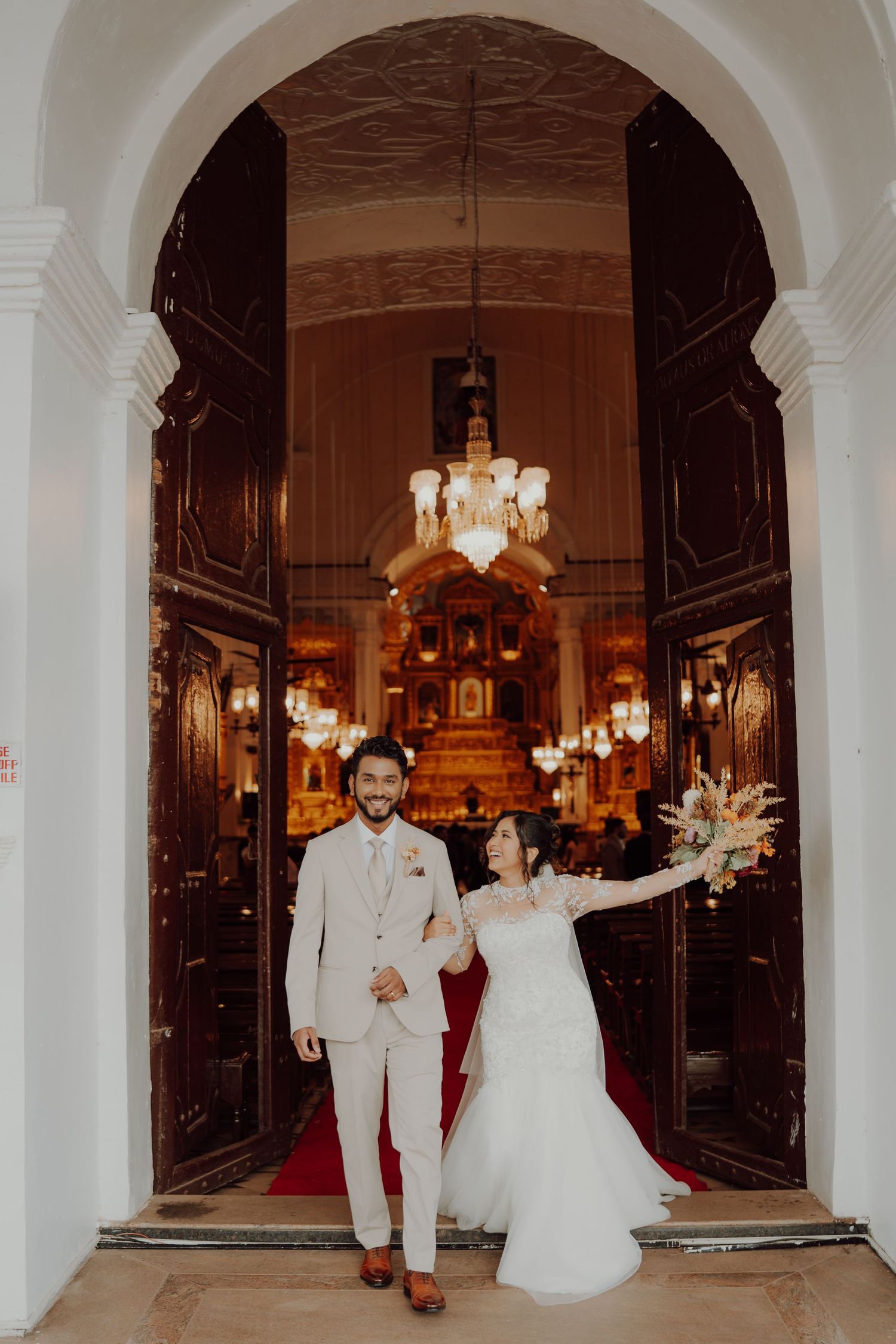 Bride and groom walk hand in hand through a grand arched church doorway as she raises her bouquet in celebration.